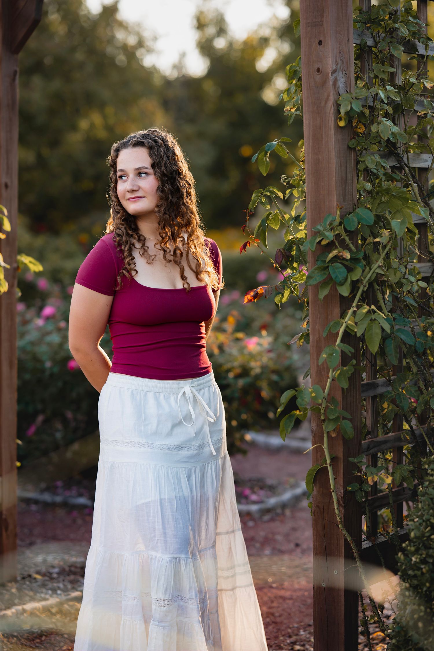 A person in a burgundy top and white skirt stands in a garden setting during golden hour lighting.