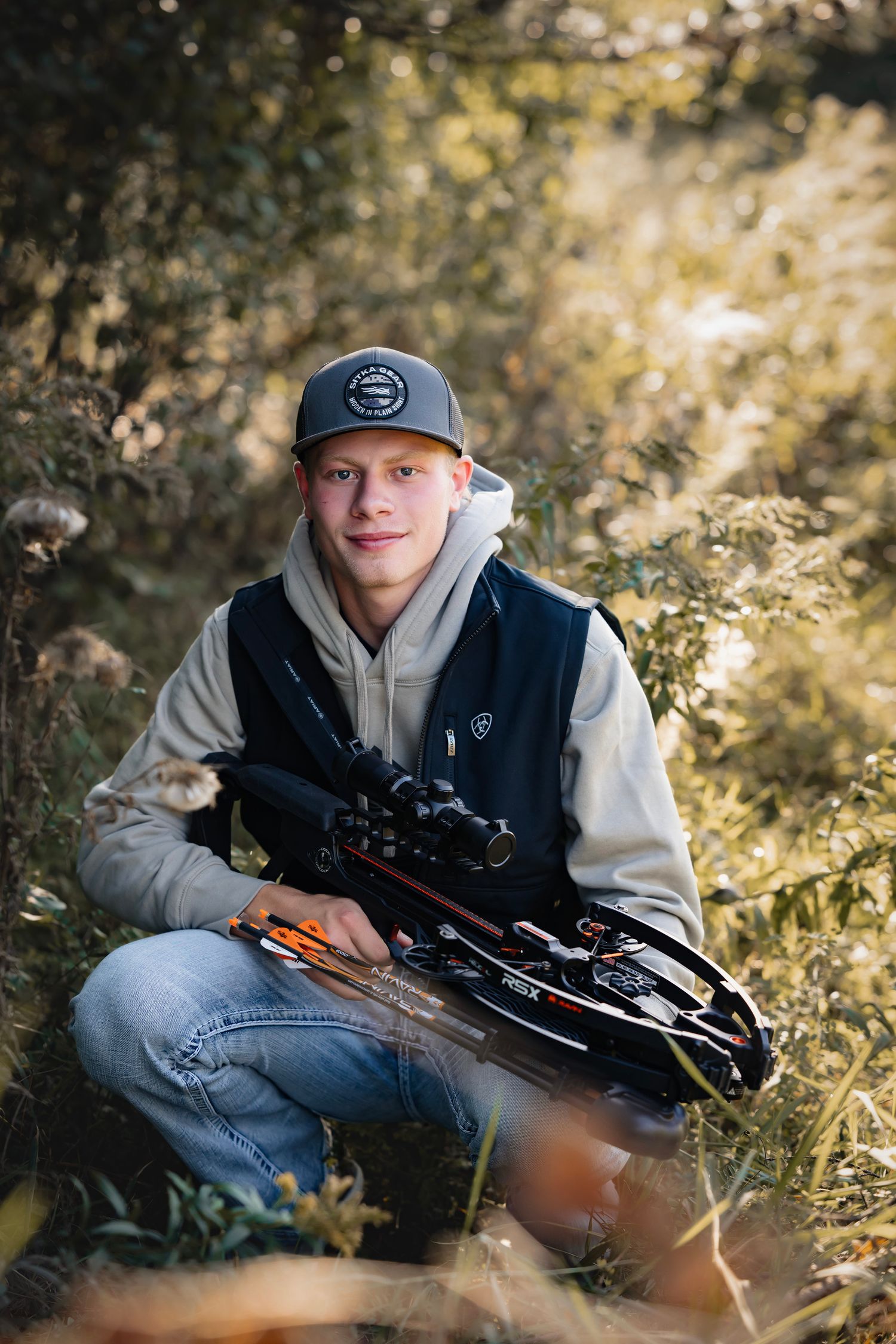 A person in a gray hoodie and baseball cap sits outdoors holding a crossbow with scope in autumn foliage.