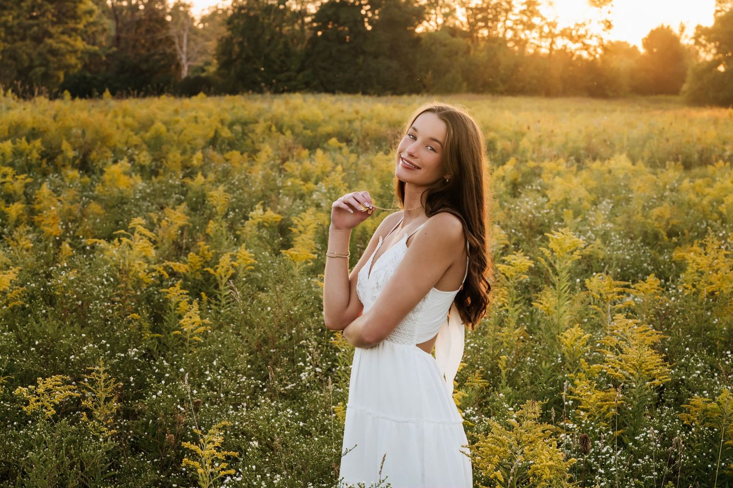 Young woman in white dress standing in golden wildflower field at sunset, smiling over shoulder.