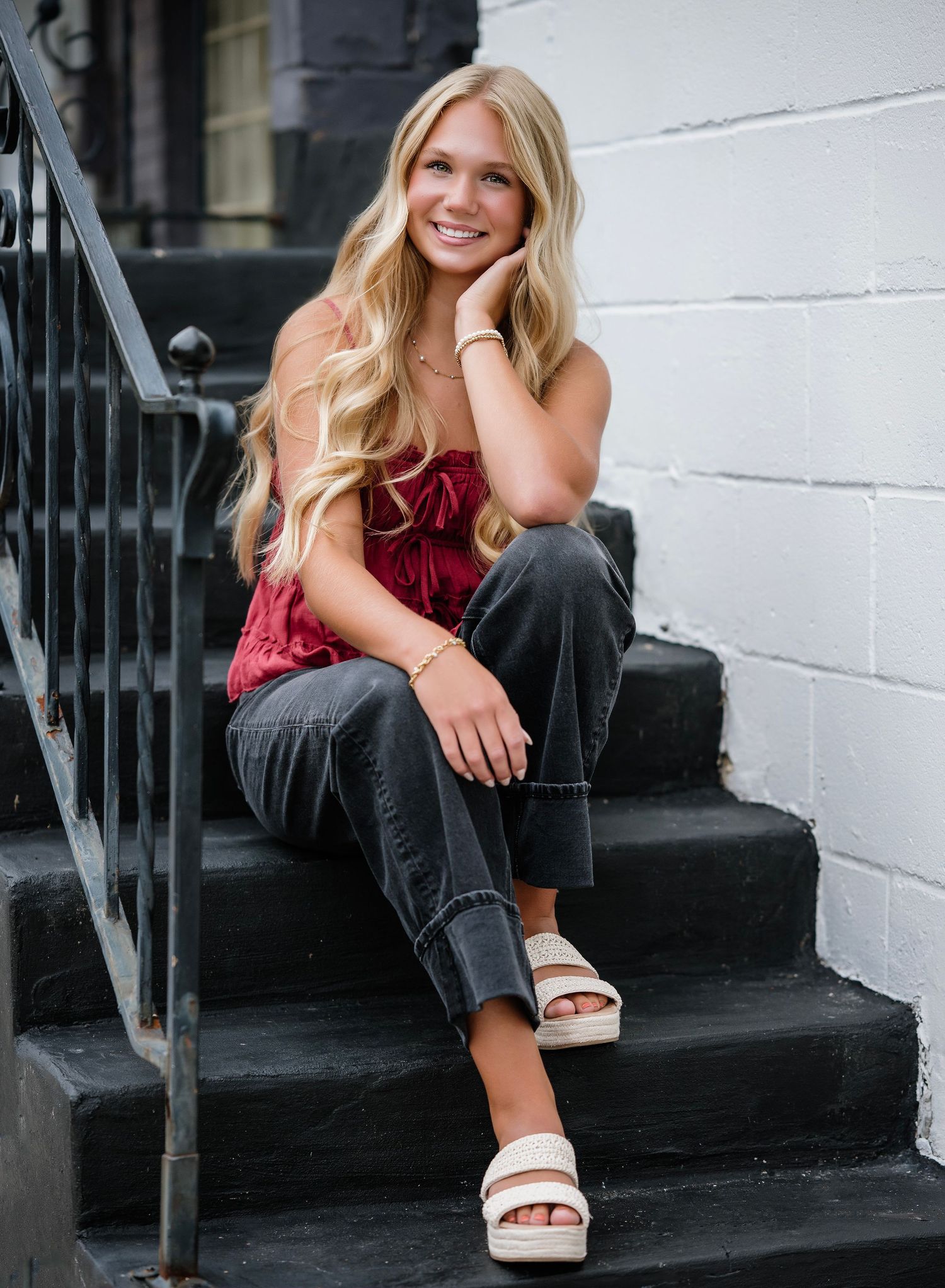 Smiling blonde teenage girl sitting on black steps wearing red top, gray jeans, and white platform sandals.