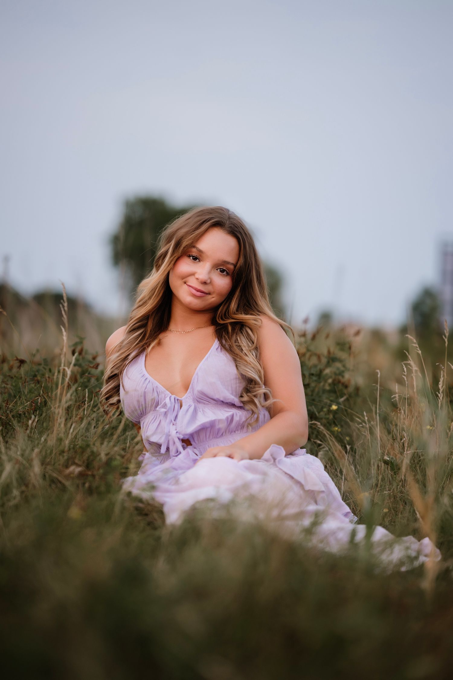 Young woman in lavender dress sitting in tall grass field, smiling softly at camera during golden hour.