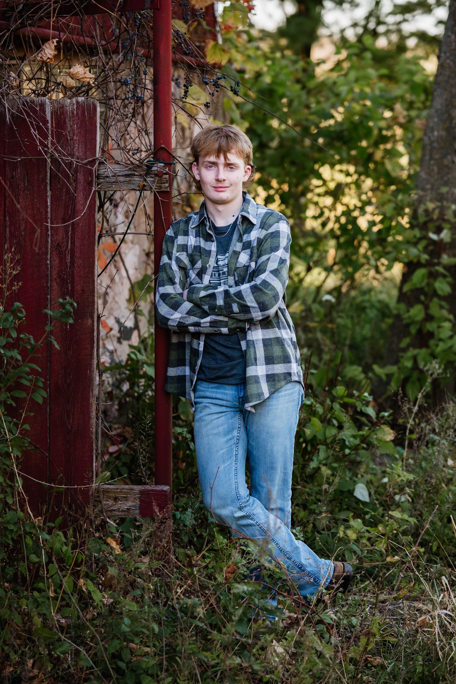 Person in plaid flannel shirt and jeans poses casually against rustic red door frame in outdoor autumn setting.