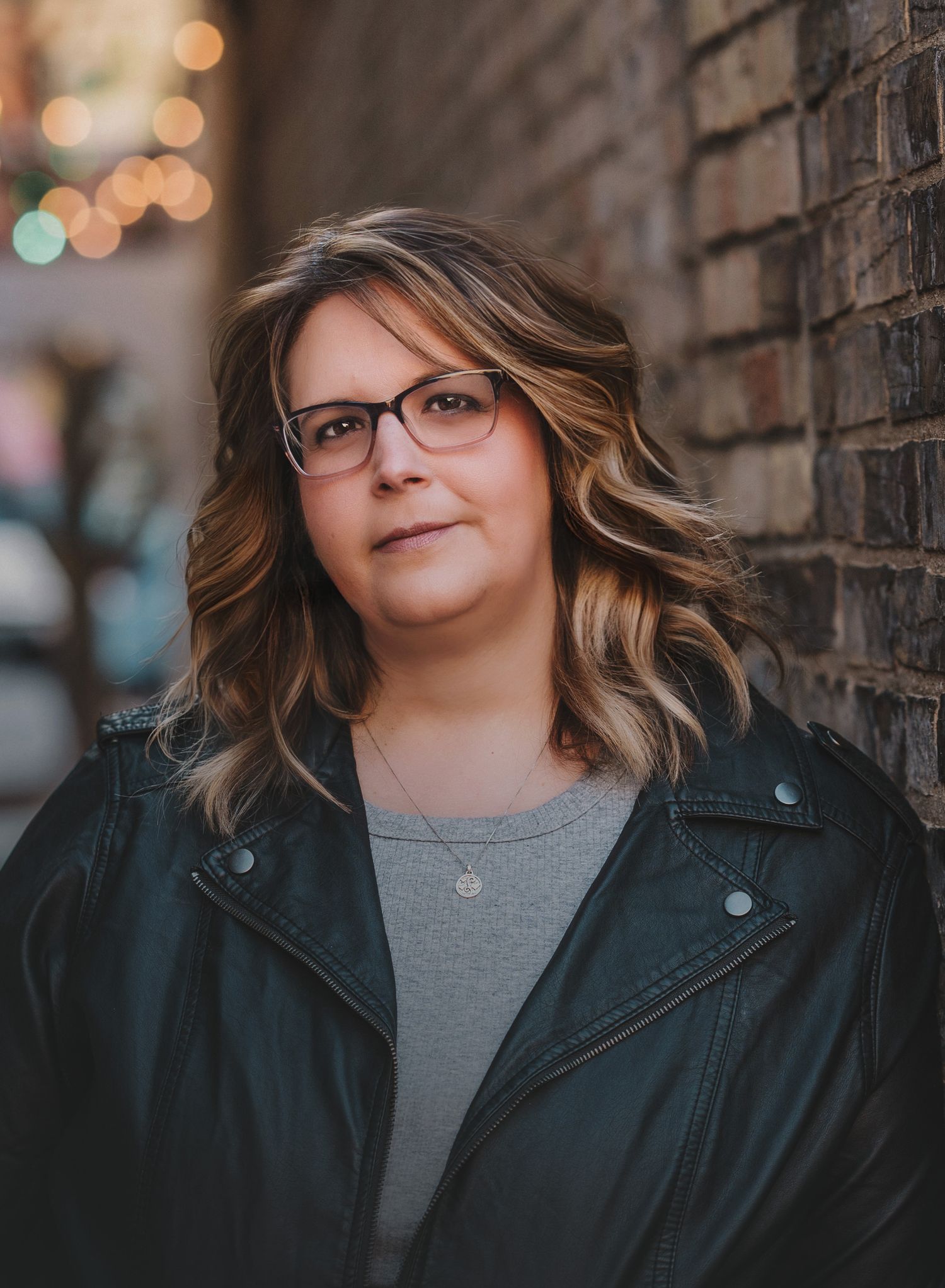 Woman with glasses and wavy hair wearing a leather jacket leaning against a brick wall outdoors.