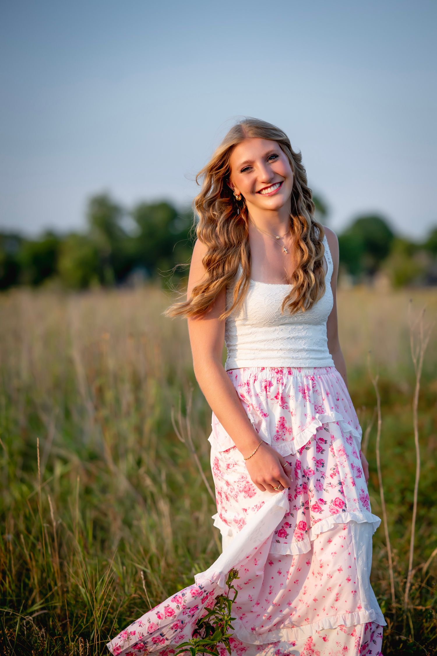 Smiling teenage girl in white top and floral skirt standing in a sunlit field with trees in the background.