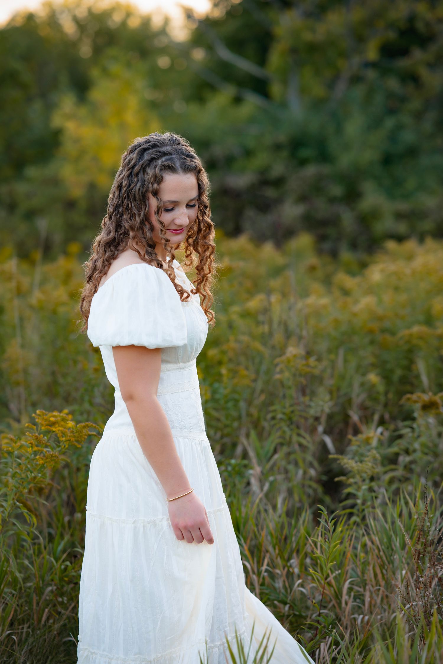 A person in a flowing white dress poses thoughtfully among tall wildflowers and grass at golden hour.