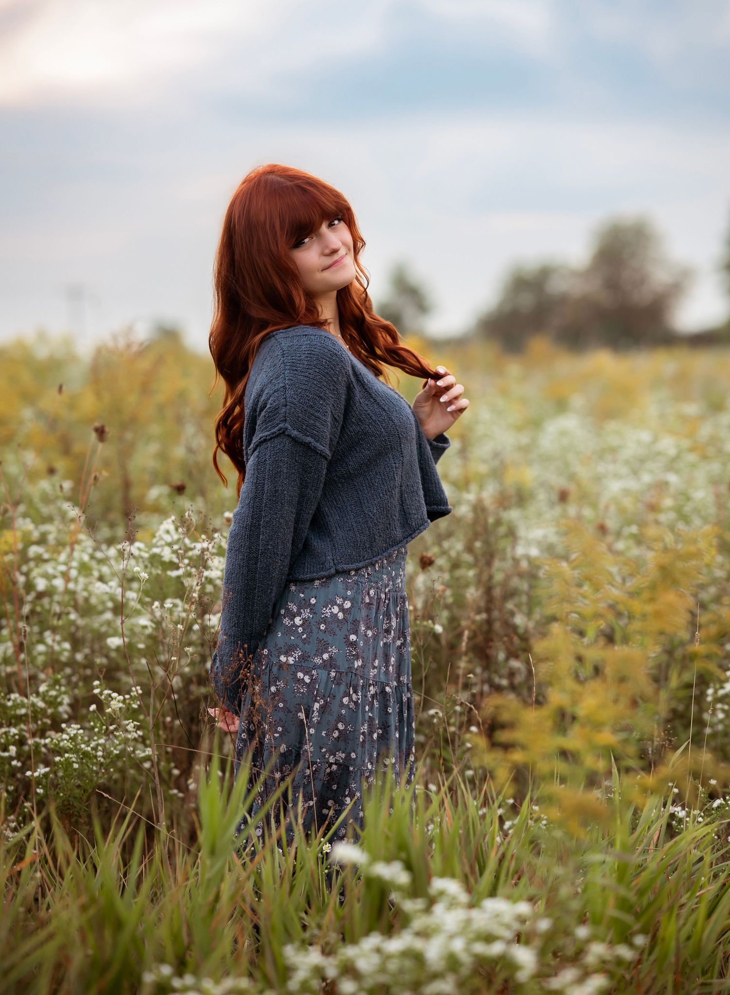A person wearing a gray sweater twirls joyfully in a meadow filled with delicate white wildflowers on a cloudy day.