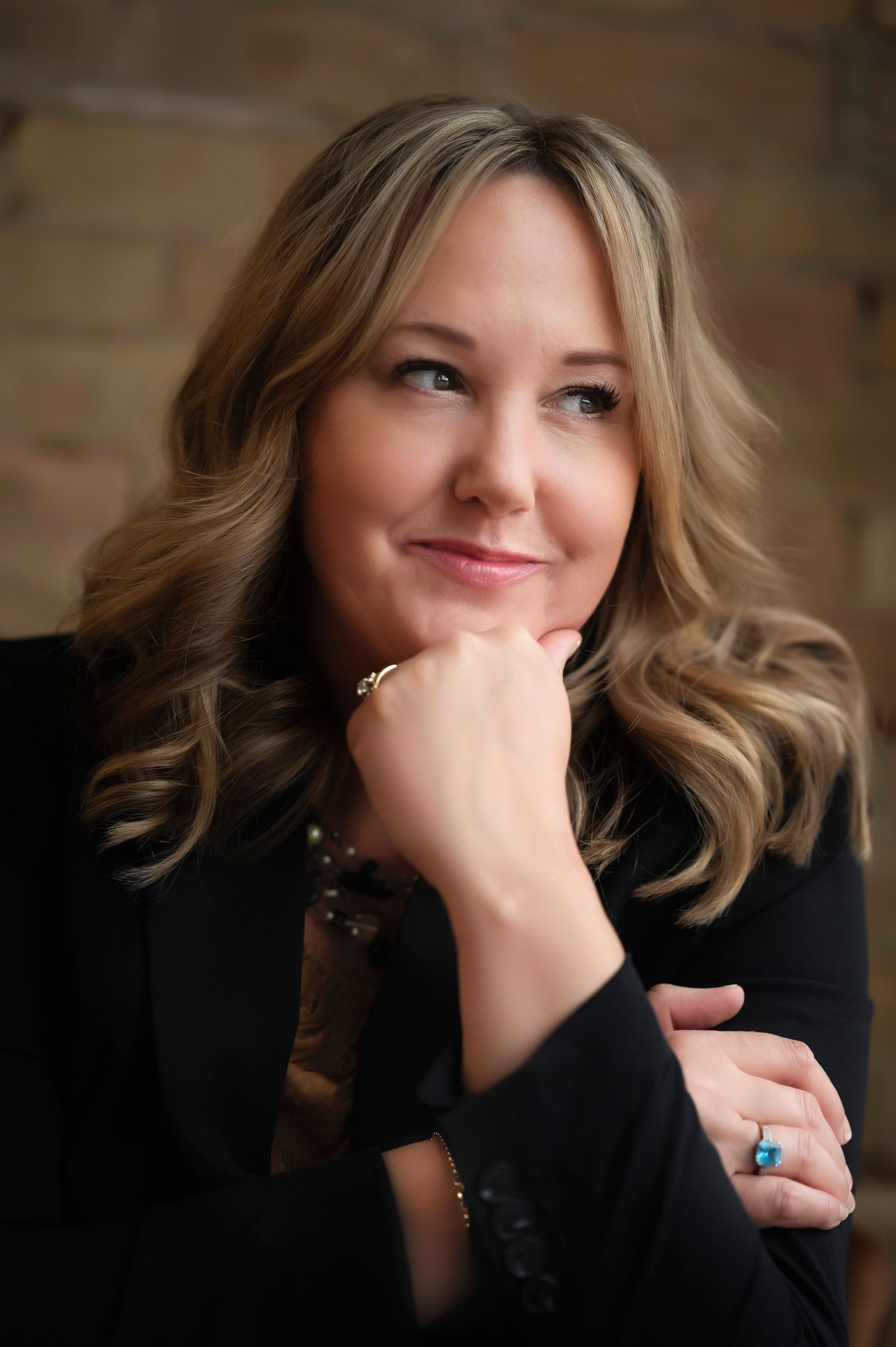 Professional portrait photo showing a blonde woman in a black blazer posing thoughtfully with hand under chin.