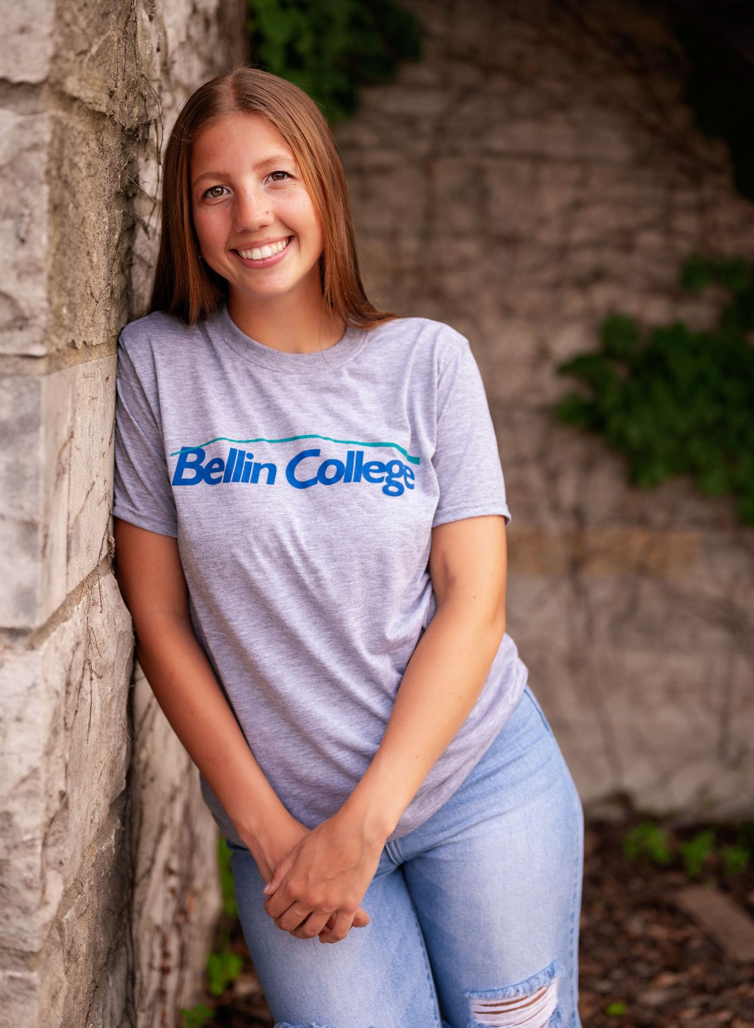 A student in a Bellin College t-shirt poses against a stone wall in an outdoor portrait series.