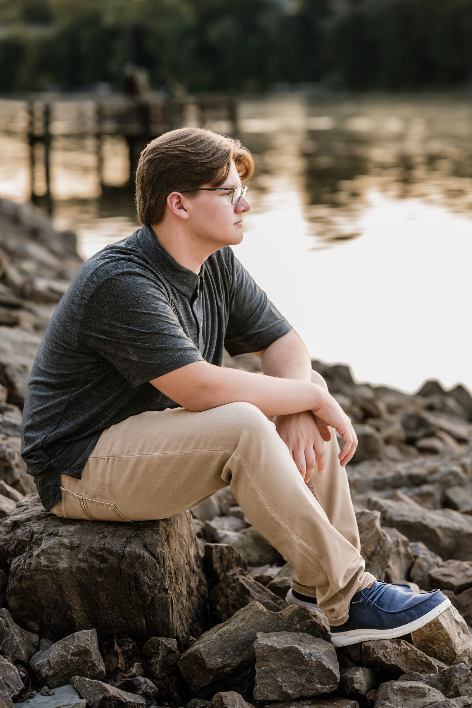 A person in a dark shirt and khaki pants sits contemplatively on rocky terrain near a reflective body of water at sunset.