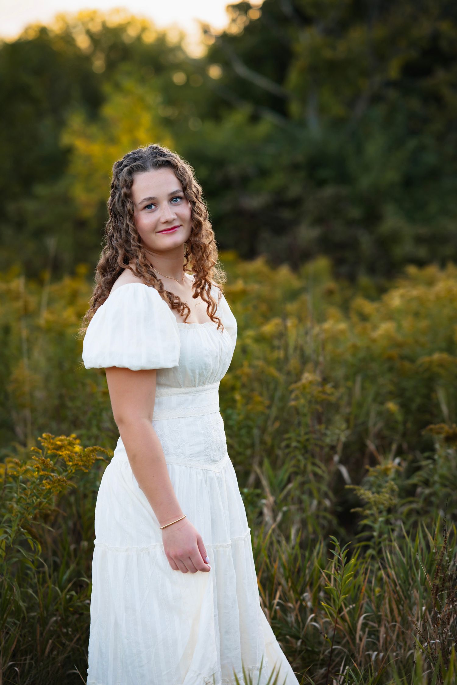 A person in a white lace dress poses in a natural outdoor setting with tall grass and golden sunlight.