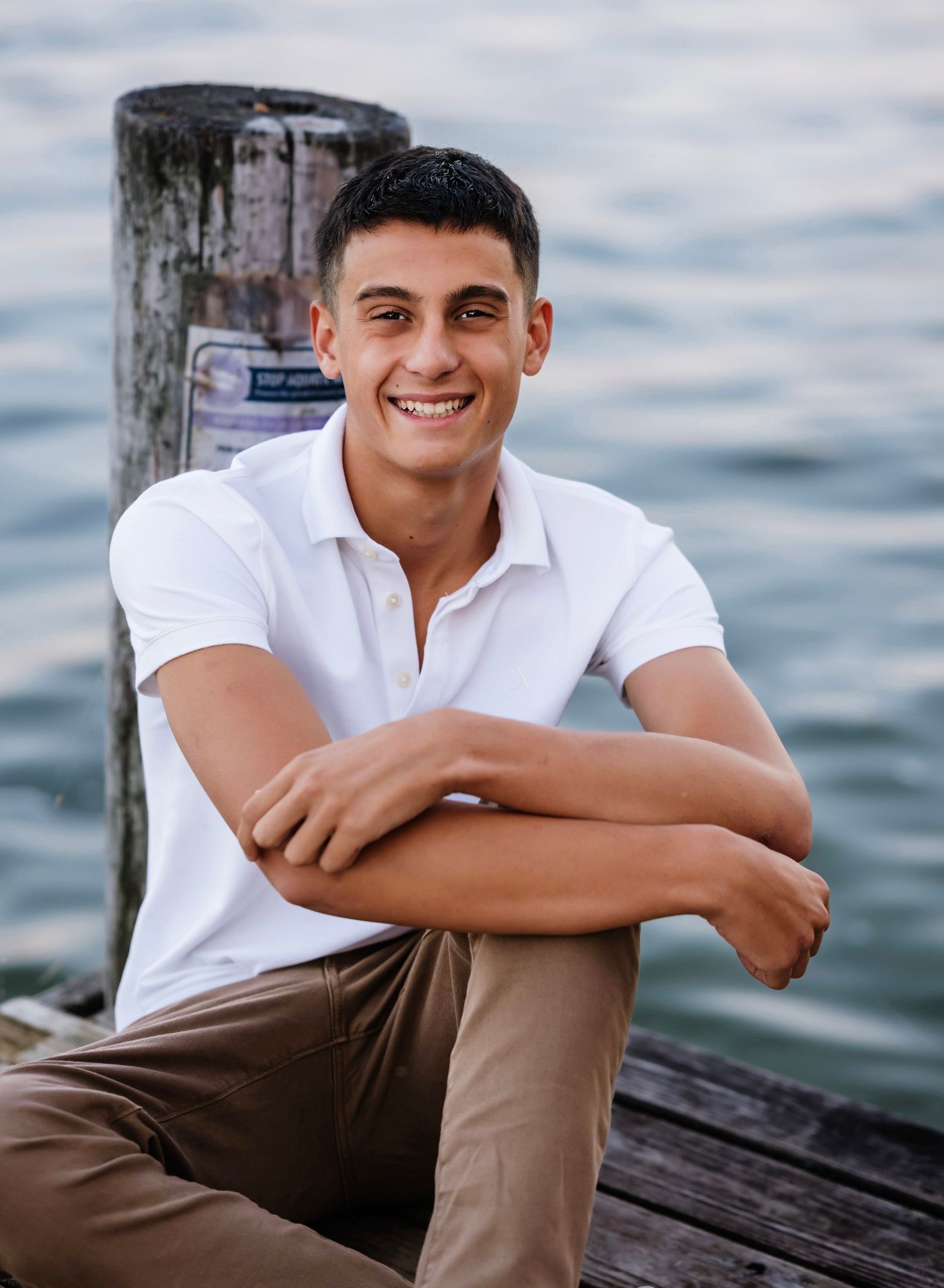 Person in white shirt and khaki pants sits casually on wooden dock by water on cloudy day.