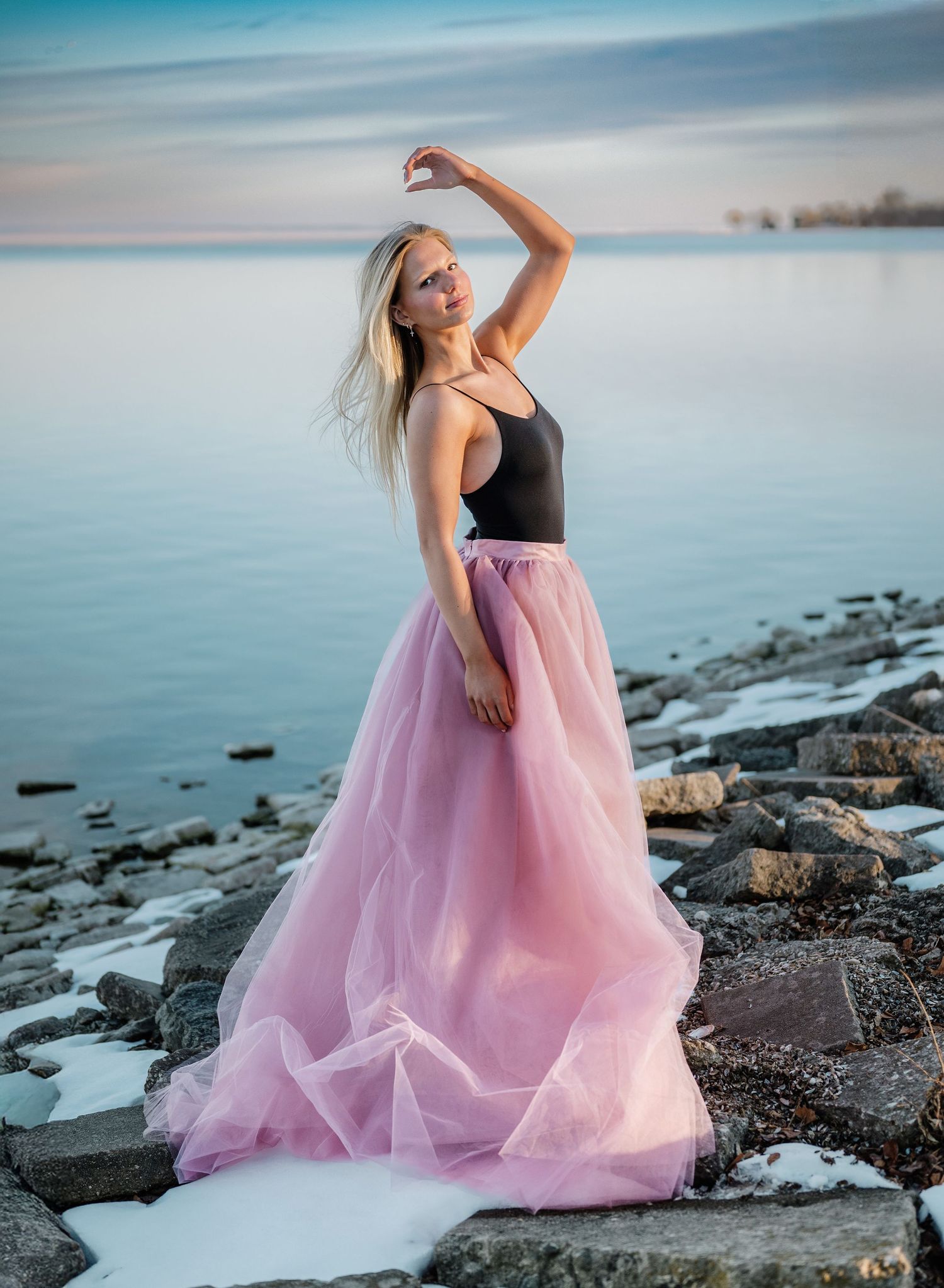Blonde woman in black top and pink tulle skirt posing on rocky shoreline at dusk.