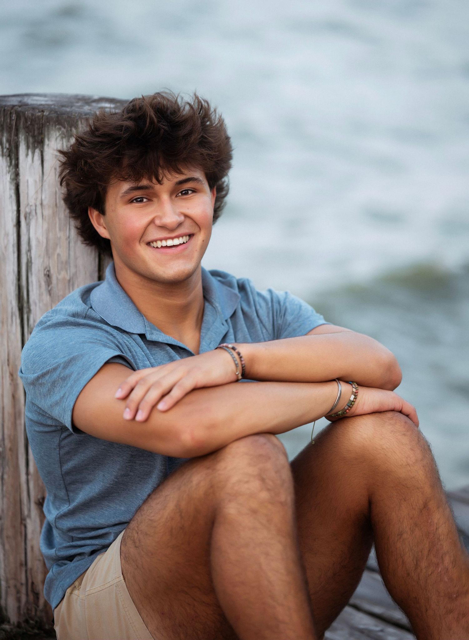 Someone sitting casually on a wooden dock by the water while wearing khaki shorts and a blue t-shirt.