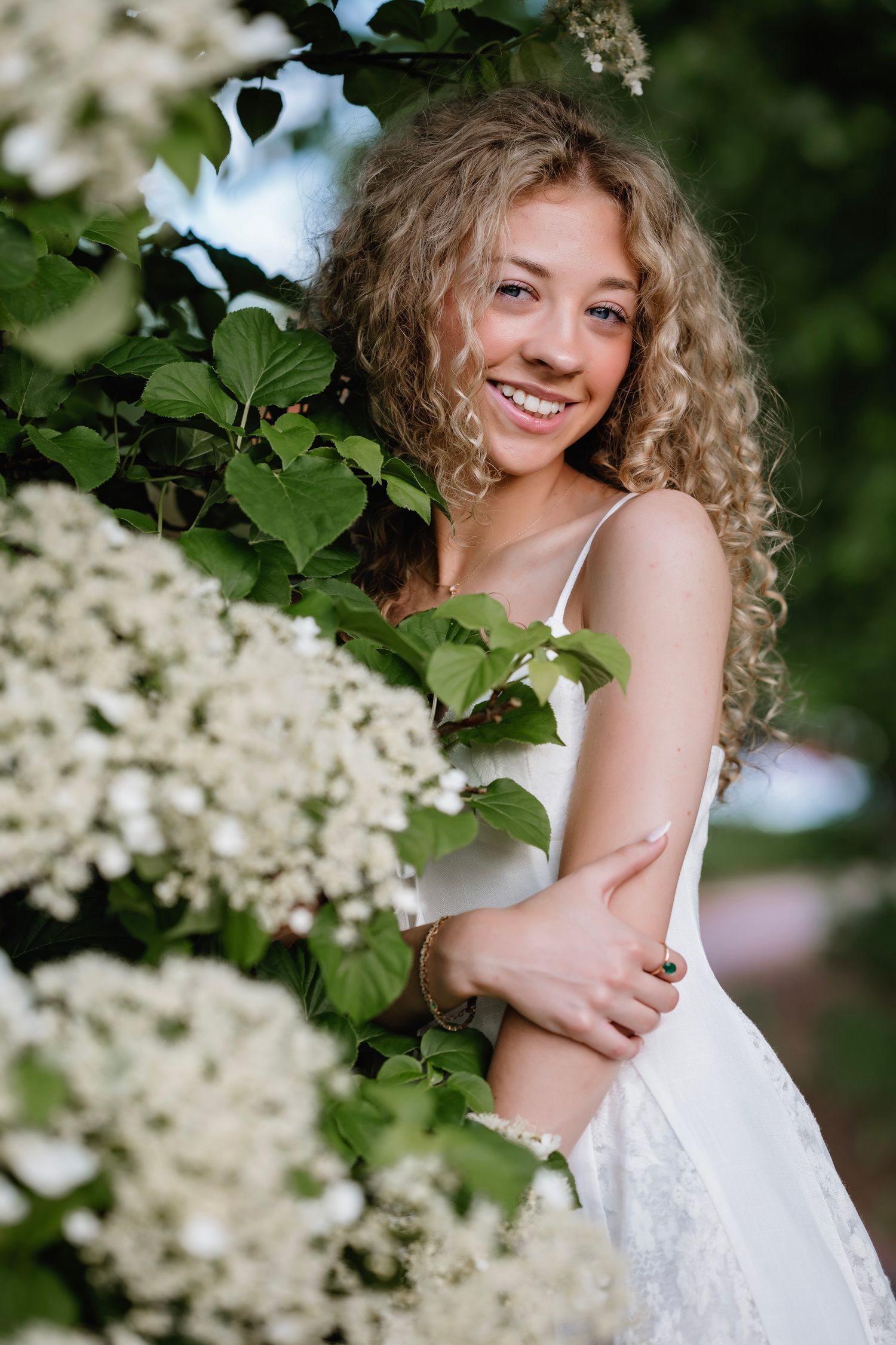 Young woman with curly blonde hair smiling in white dress among blooming white flowers and green leaves outdoors.