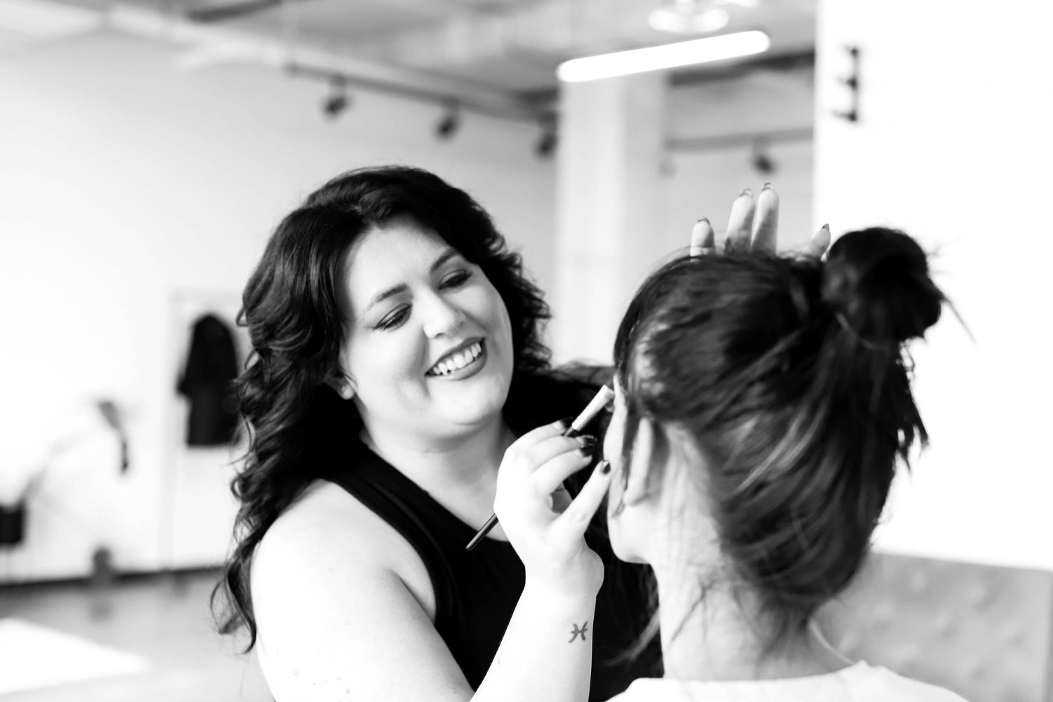 Black and white photo of a smiling makeup artist applying makeup to a client in a bright studio.