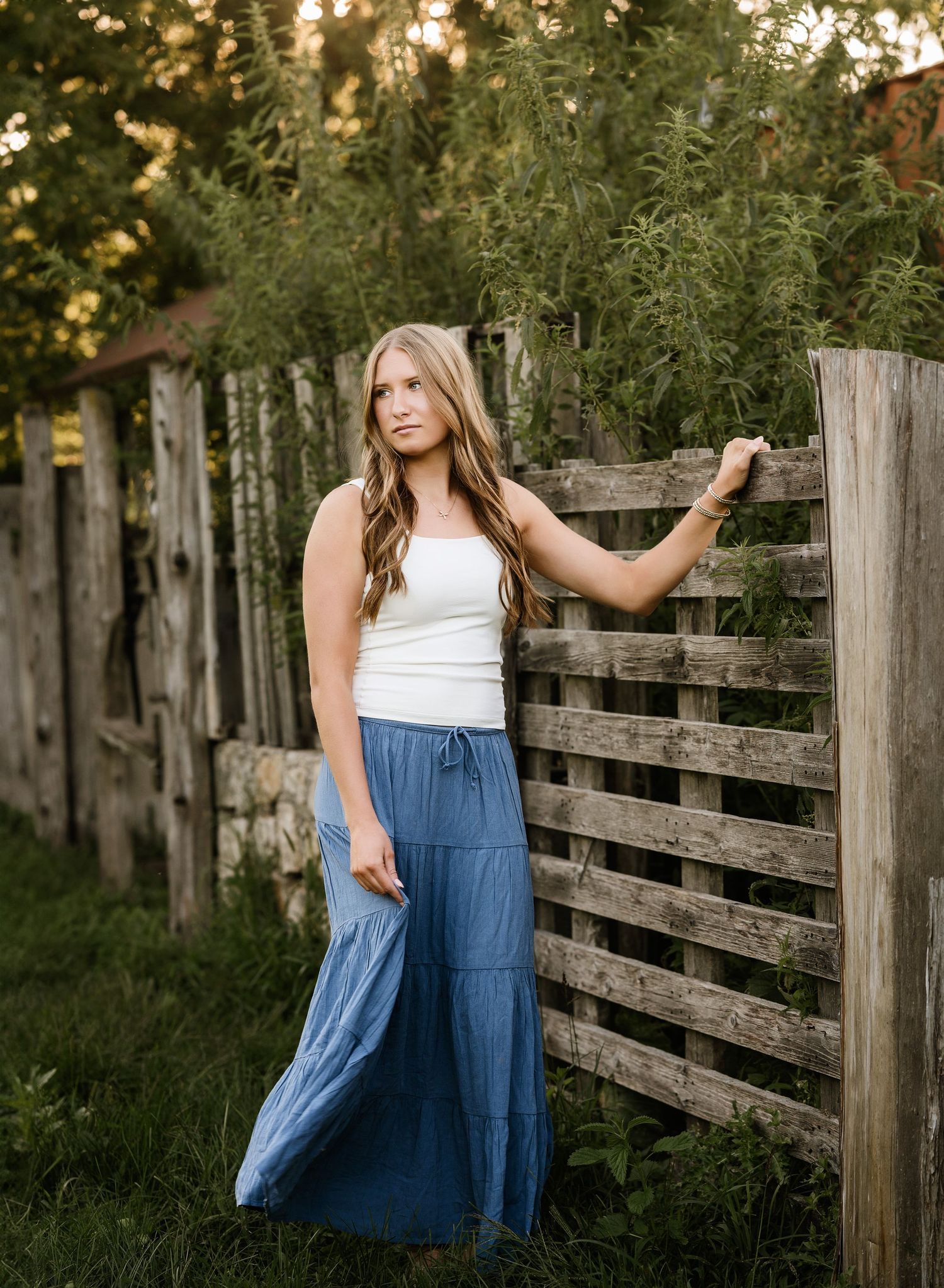 Young woman in white top and blue maxi skirt leaning against rustic wooden fence with lush greenery backdrop.