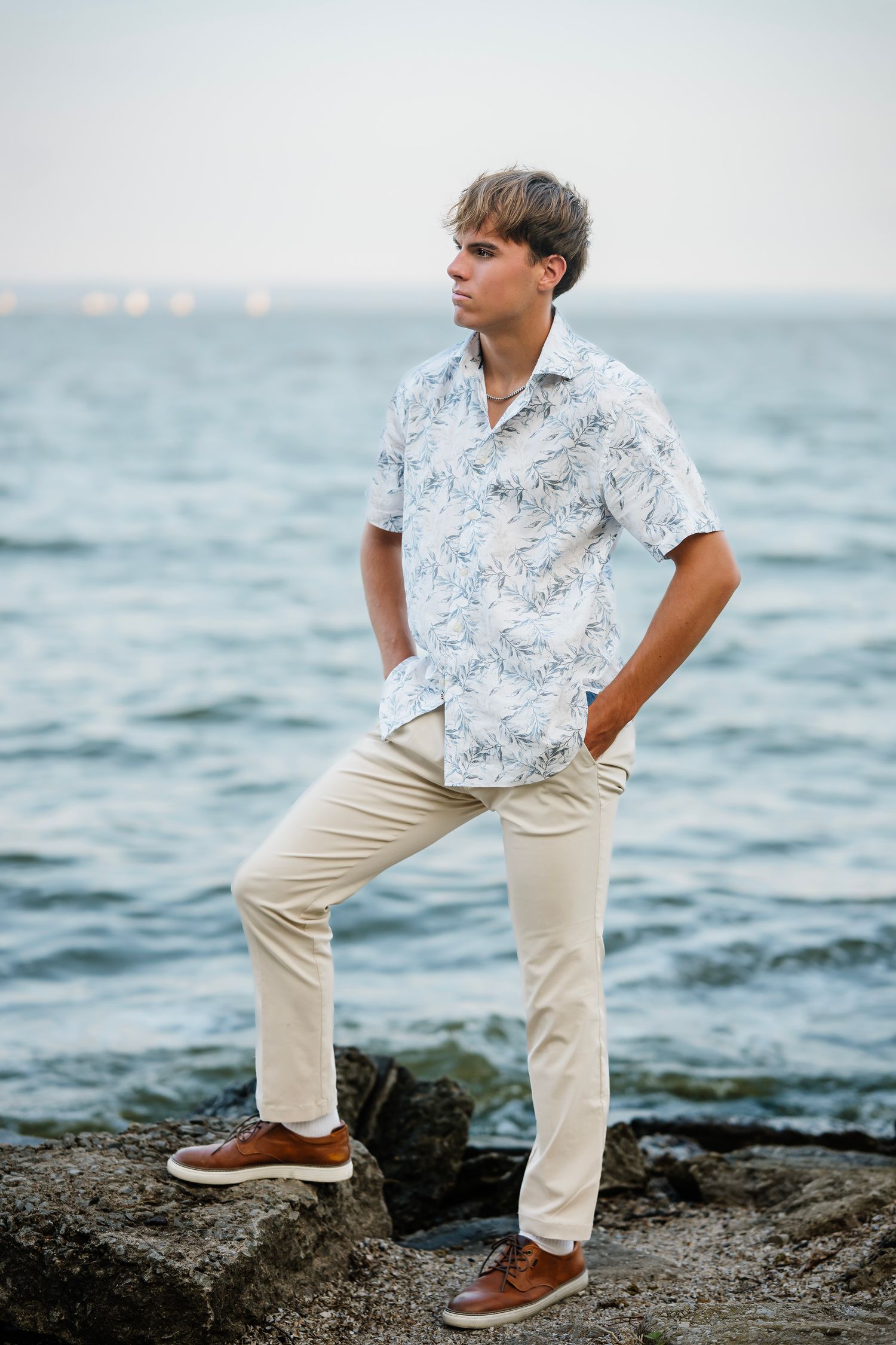 Person in white floral shirt and khaki pants poses on rocky coastal shoreline with ocean waves in background.