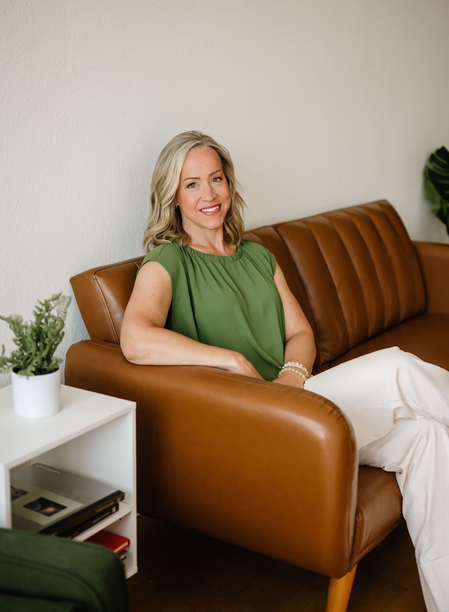 Someone sits comfortably on a brown leather couch wearing a sleeveless green top with indoor plants and white decor visible.