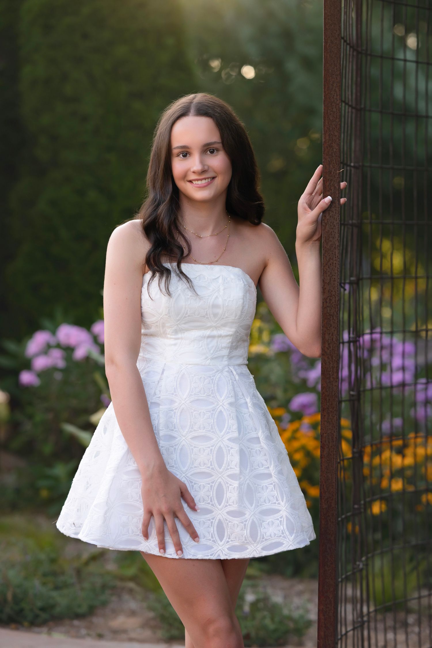 Young woman in white strapless dress posing by metal fence with colorful garden flowers in background.