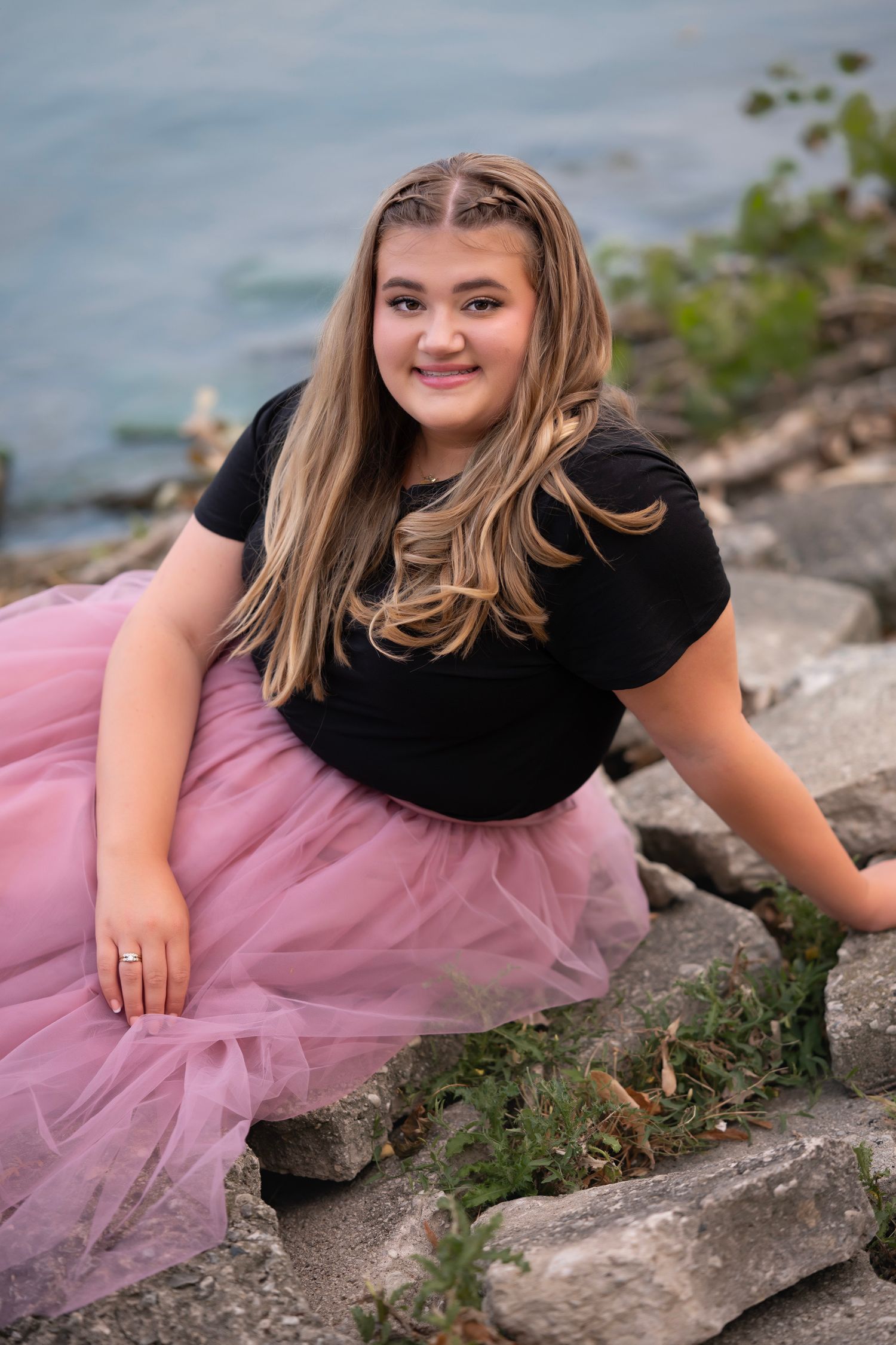 Teen girl in black top and pink tulle skirt sitting on rocks near water, smiling for a portrait photo.