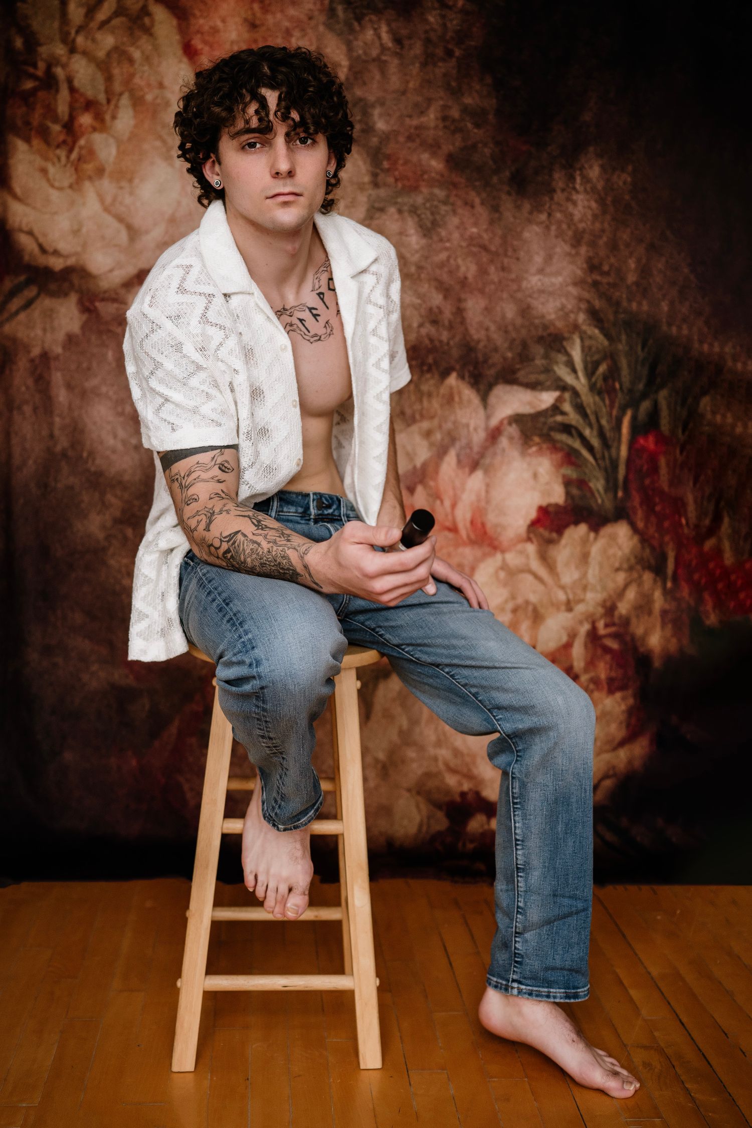 Young man with curly hair and tattoos sits on wooden stool, wearing open white shirt and jeans, floral backdrop.