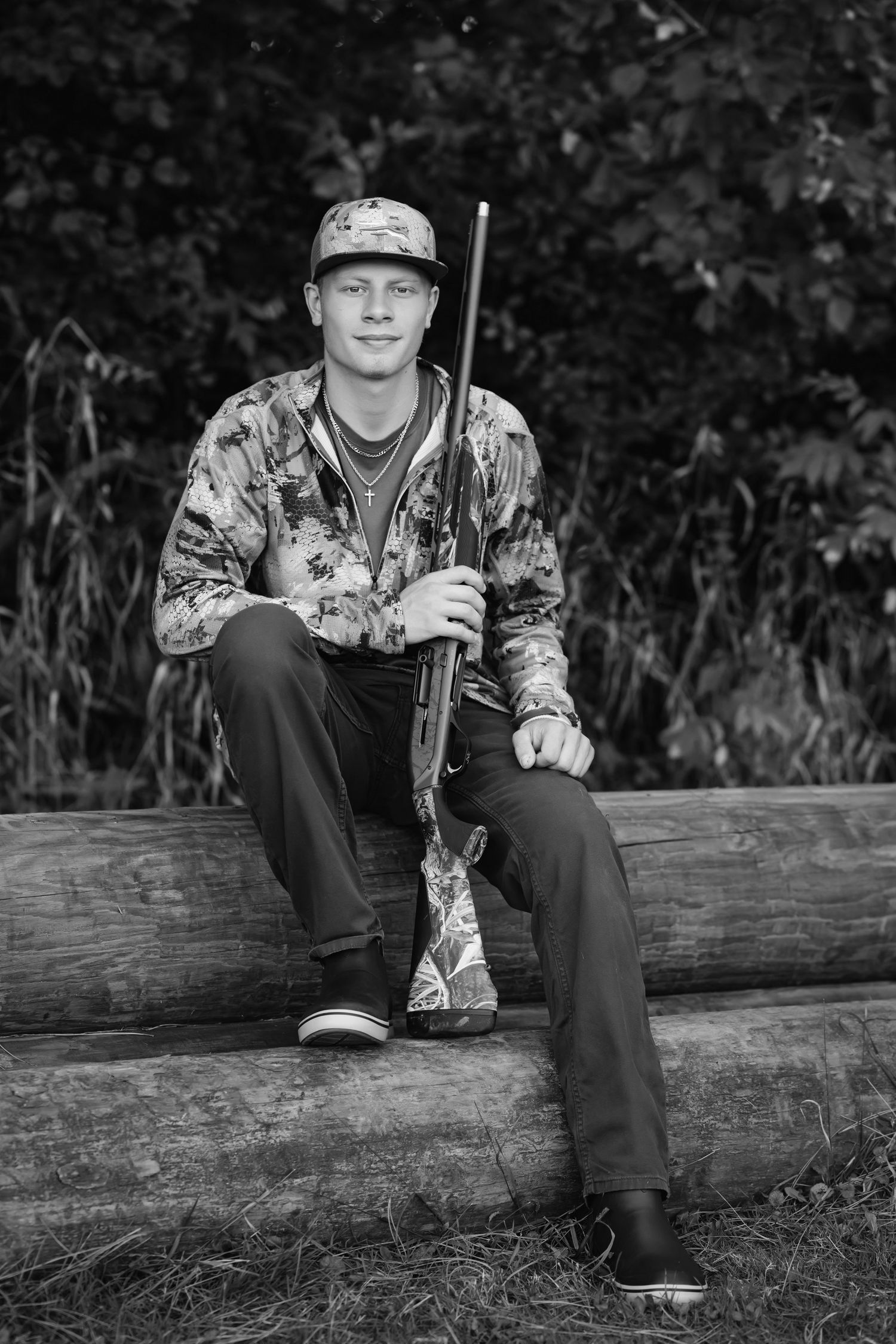 Young hunter in camo jacket and cap sits on log holding rifle in black and white outdoor portrait.