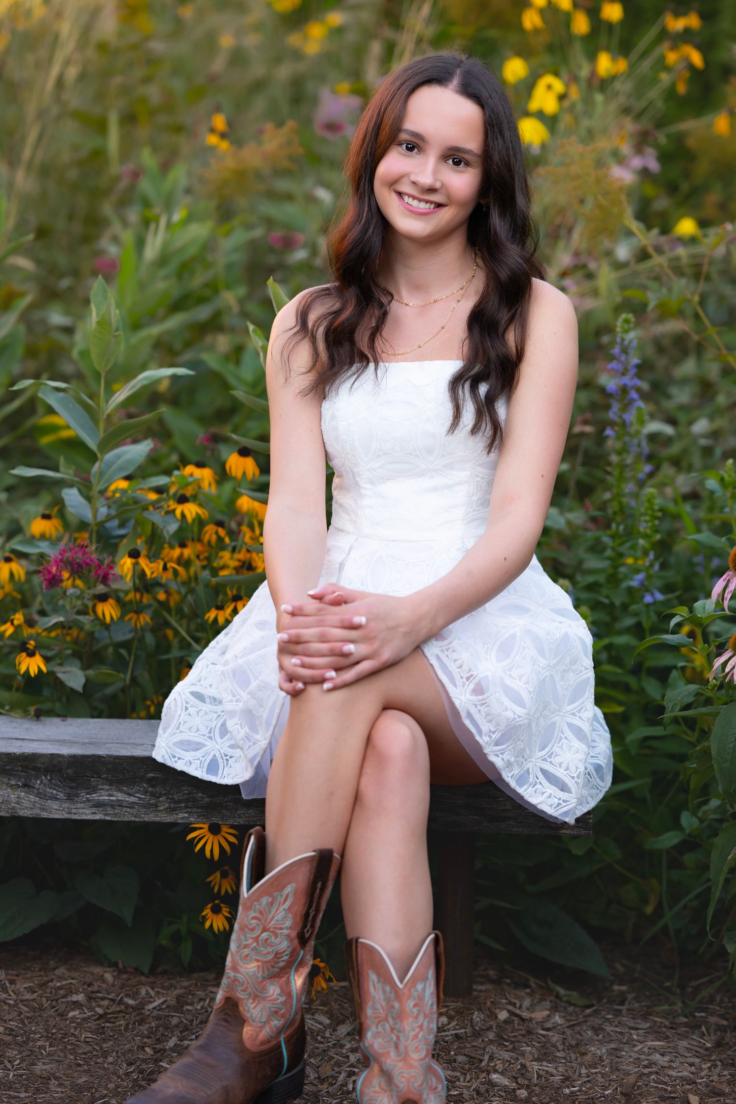 Young woman in white lace dress and cowboy boots sitting on bench surrounded by yellow wildflowers.