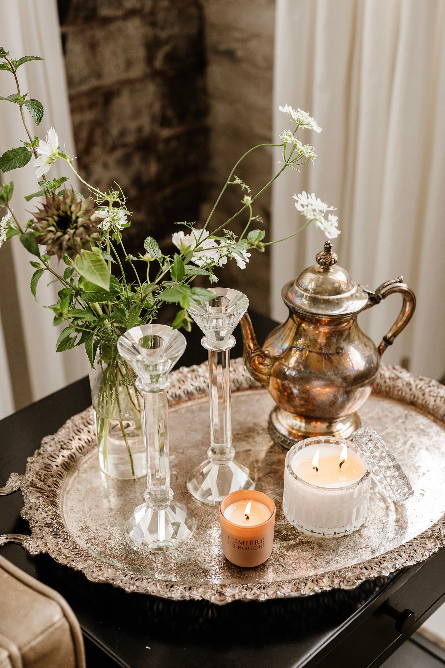 Elegant silver tray with candles, crystal candlesticks, antique teapot, and fresh wildflowers arrangement on dark table.