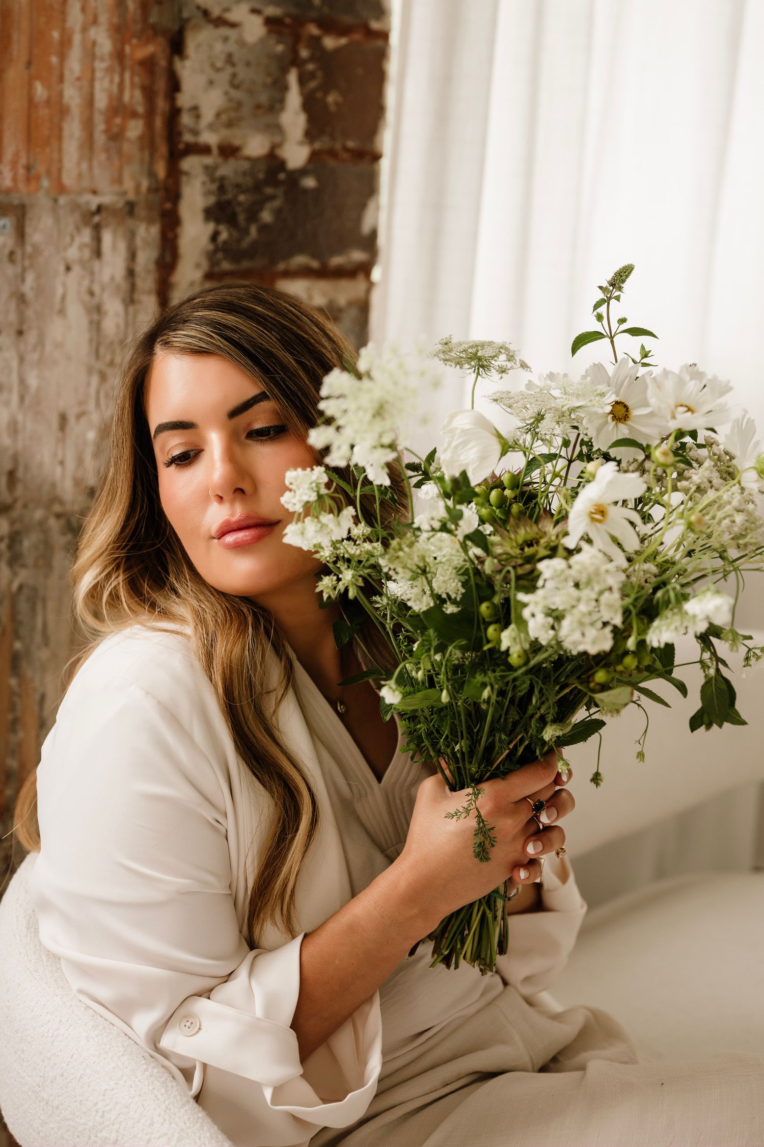 Woman in white blazer holding a bouquet of white wildflowers, posing near a window with soft natural light.