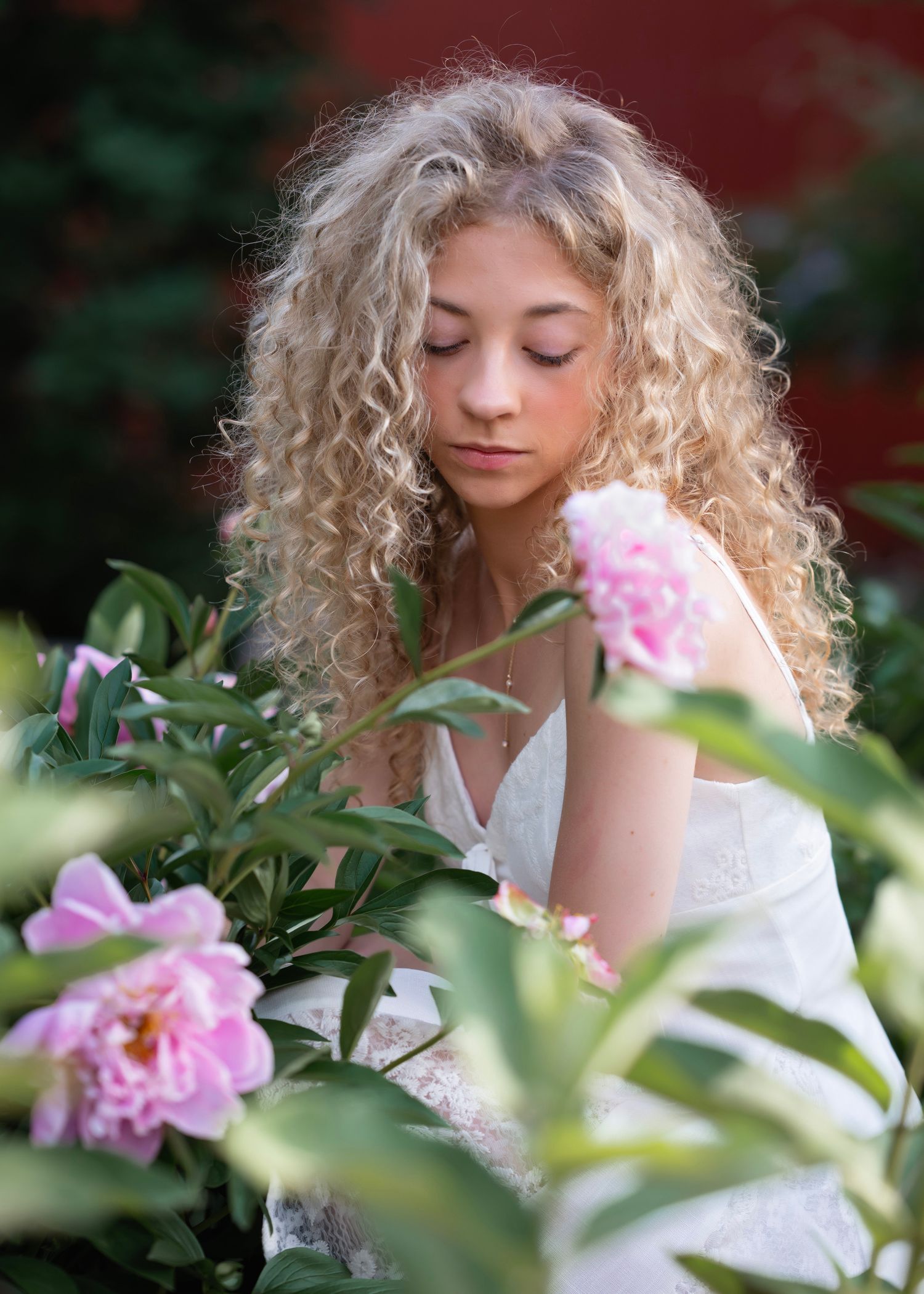 Young woman with curly blonde hair gazes down at pink peony blooms in a lush garden setting.