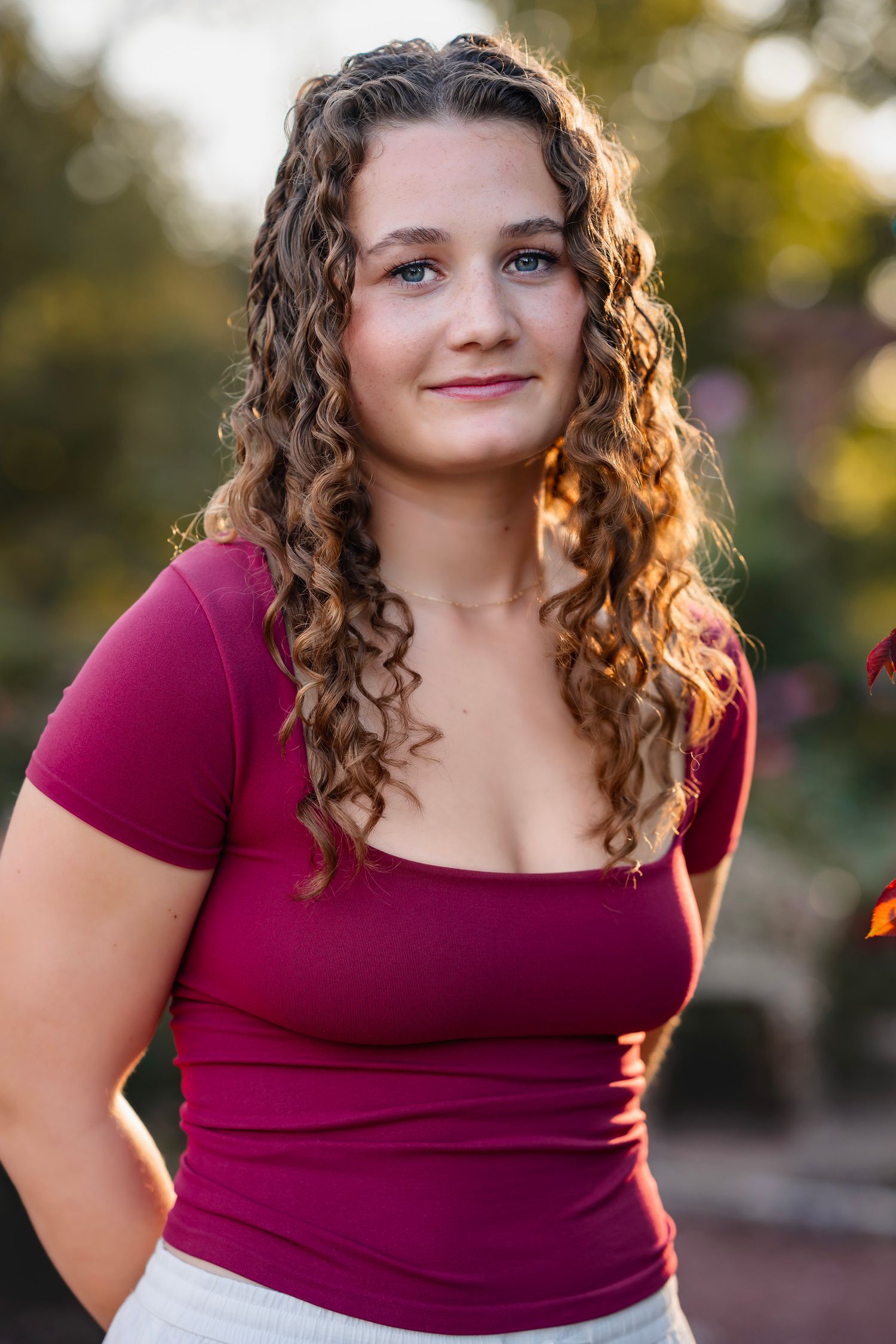 Young woman with curly hair wearing a burgundy top, posing outdoors in warm golden hour sunlight.