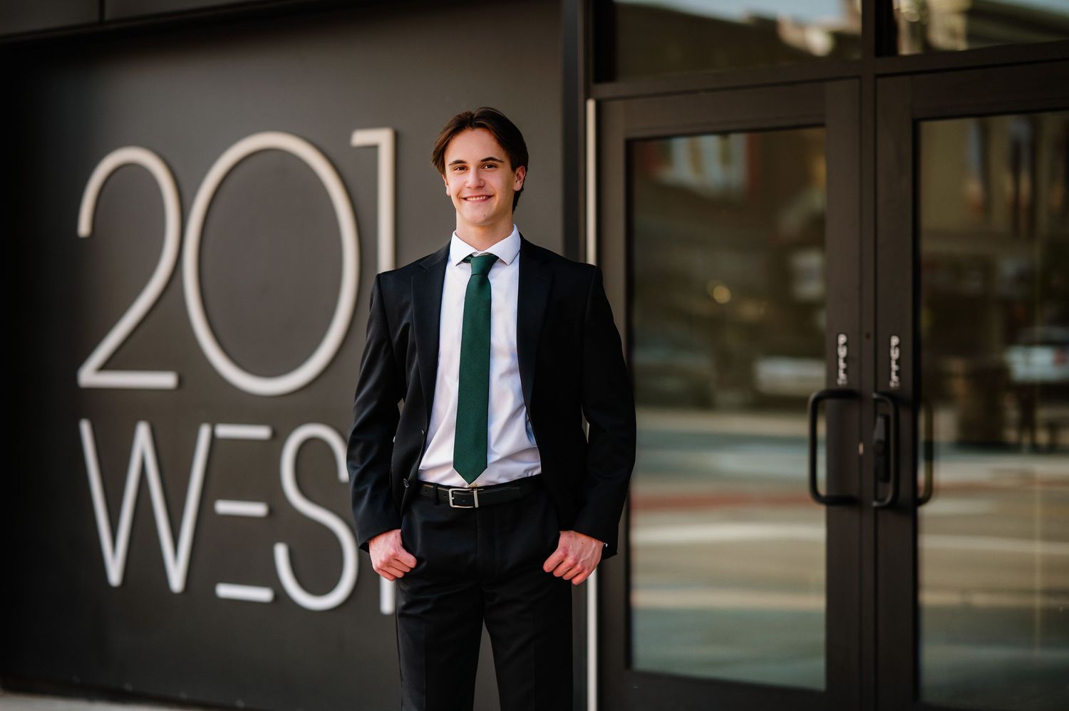 Young professional man in black suit and green tie posing outside 201 West building entrance.