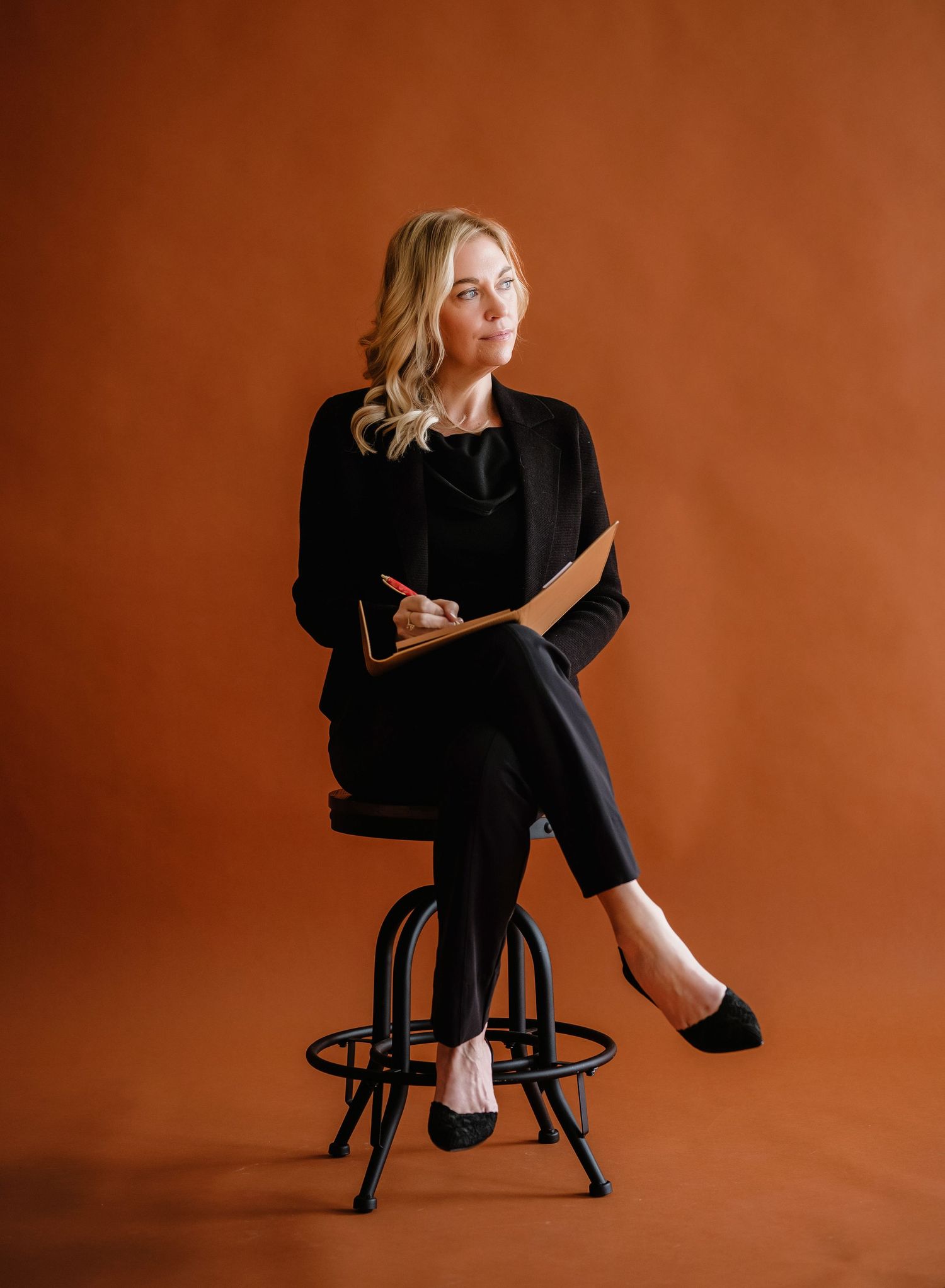 Professional portrait of someone seated on a black bar stool against a brown backdrop wearing an elegant black outfit.