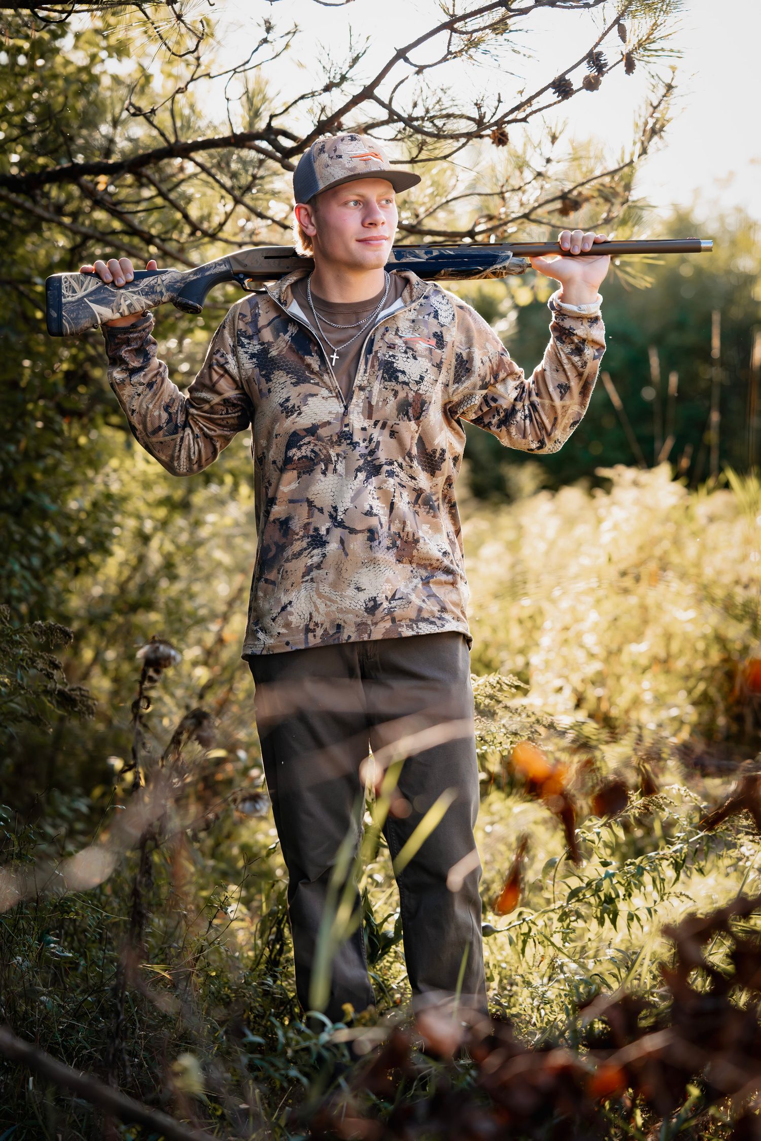 Hunter in camouflage clothing stands in nature holding a rifle over their shoulder during golden hour.