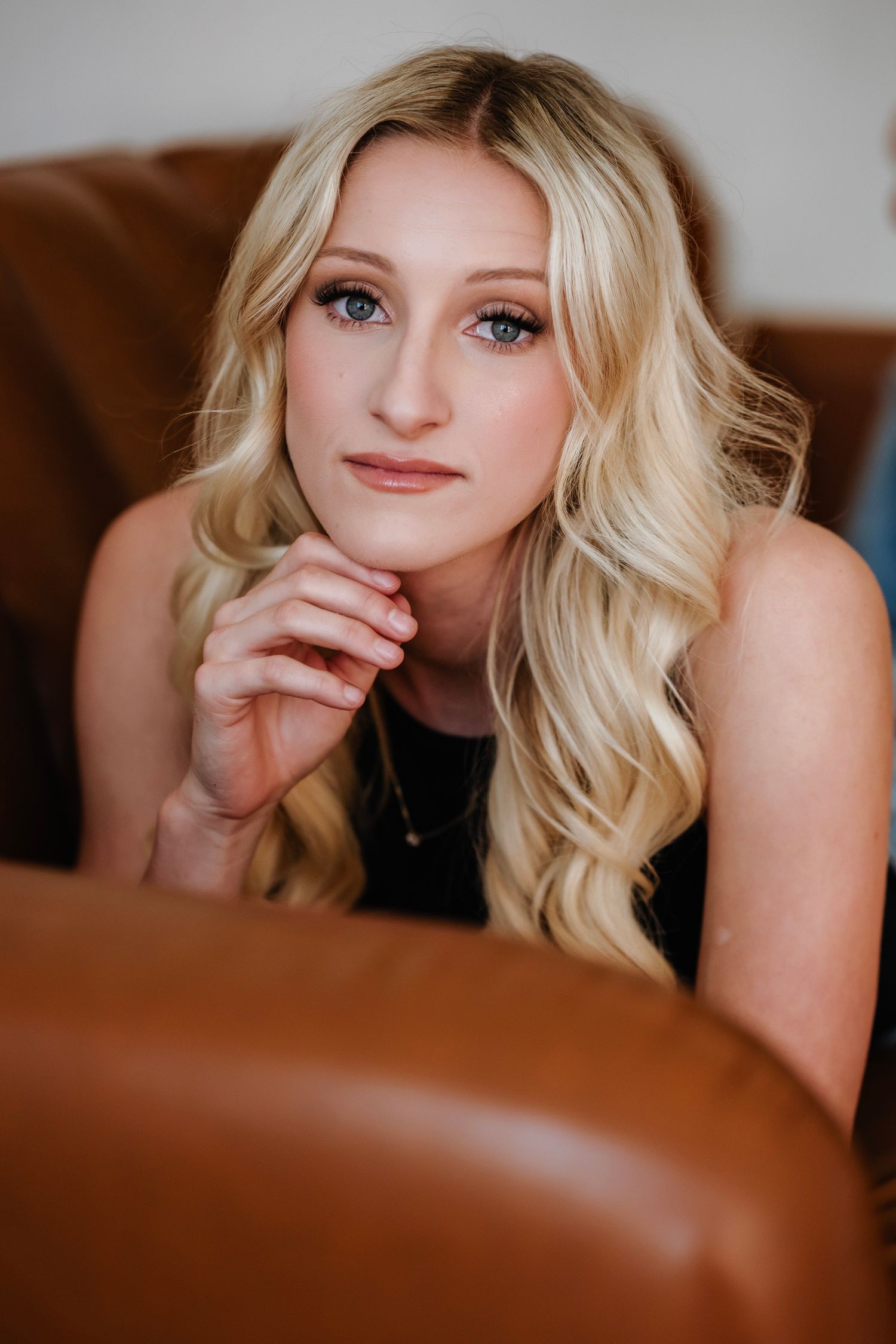 Young blonde woman with blue eyes resting chin on hand, sitting in a brown leather chair.