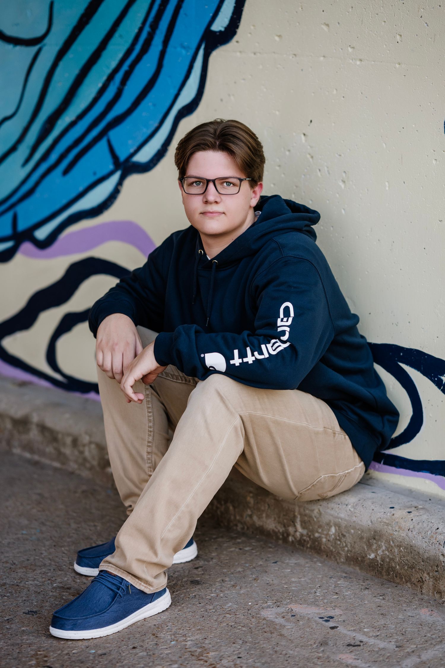 Person in a navy hoodie and khaki pants sits casually against a wall with blue street art details.