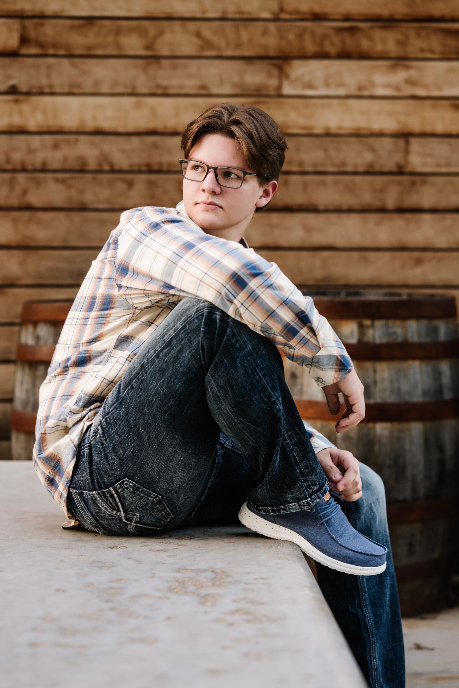 Person in plaid shirt and jeans sitting casually on wooden surface with rustic wood paneling background.