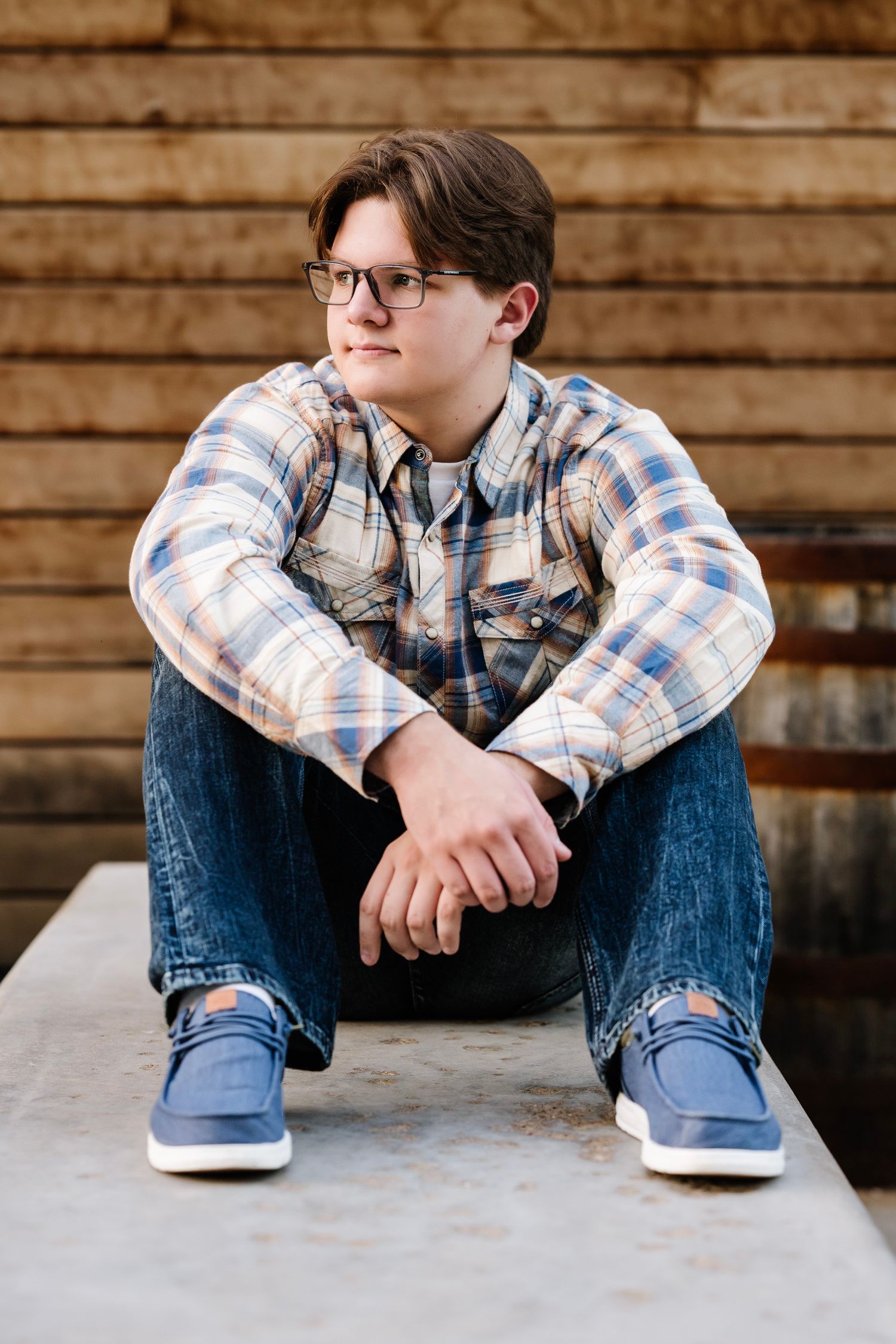 A person in a plaid shirt and jeans sits casually on a concrete surface against a rustic wooden wall background.
