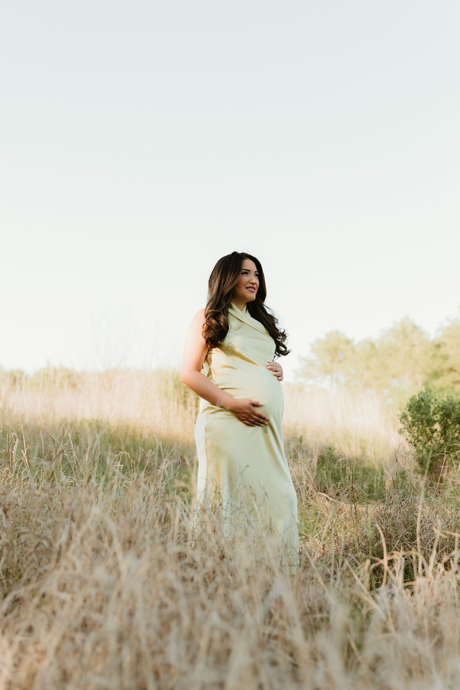 Woman in white dress poses gracefully in tall grass during sunset maternity photoshoot.