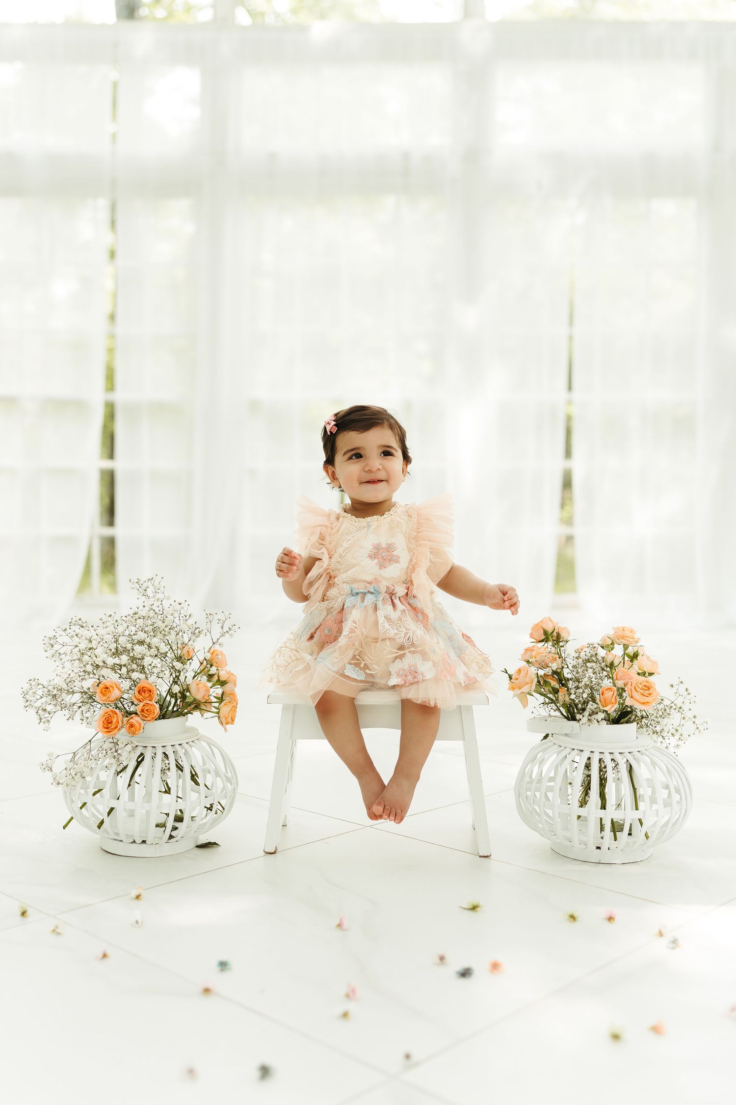 A baby in a pink dress sits between floral arrangements on a white backdrop.