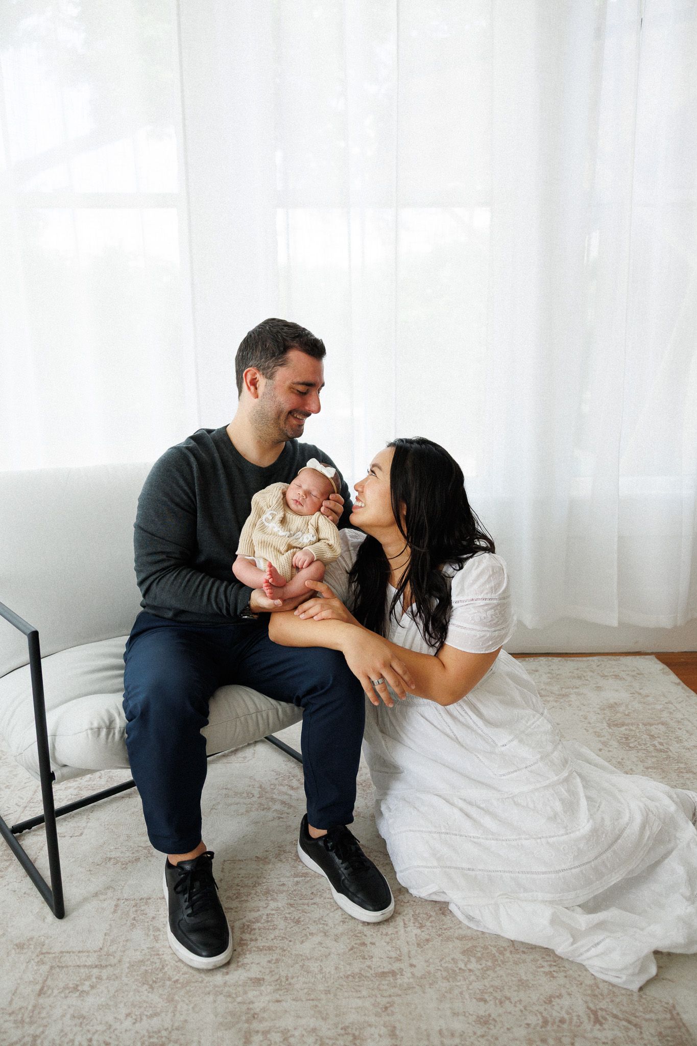 A couple smiles at their newborn baby in a bright, airy room with white curtains.