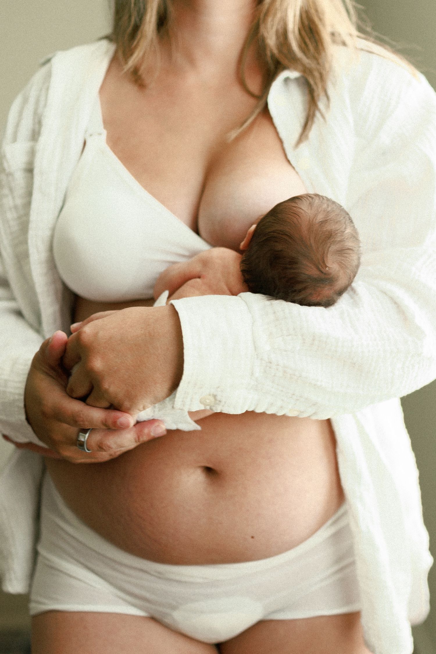 New mother breastfeeding newborn baby while showing postpartum belly, wearing white bra and underwear.