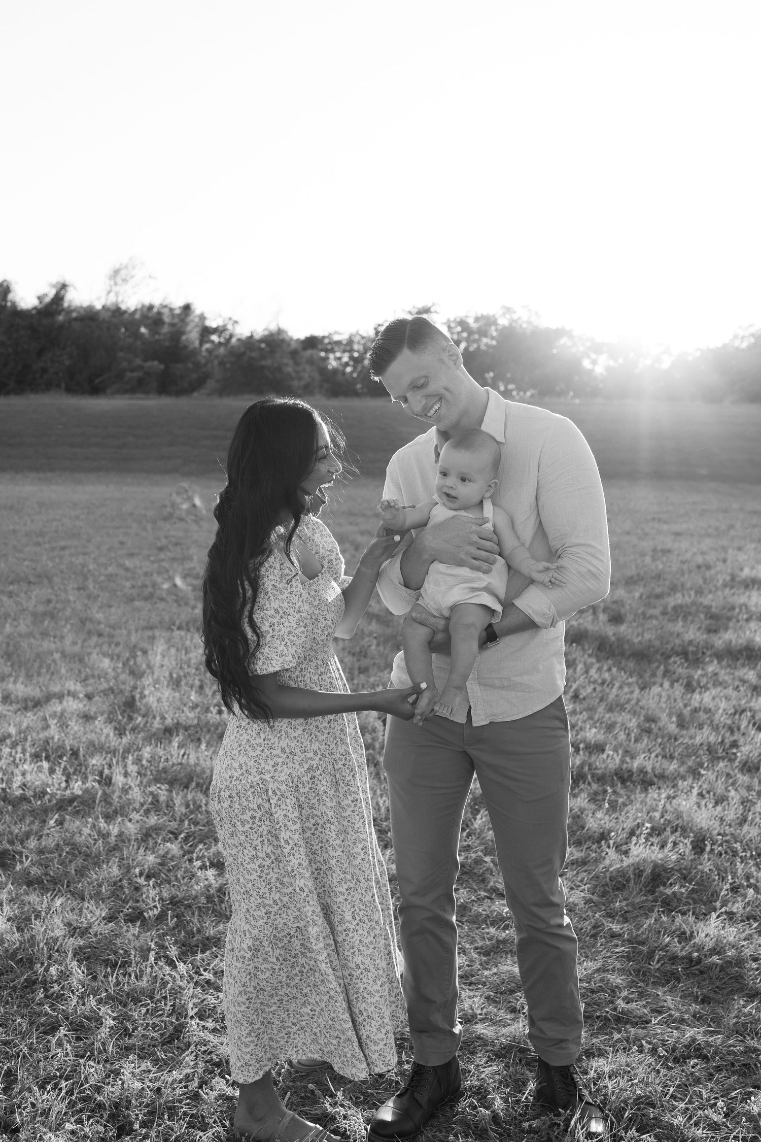 A family sharing a tender moment together in a sunlit field at sunset wearing casual and floral dress attire.