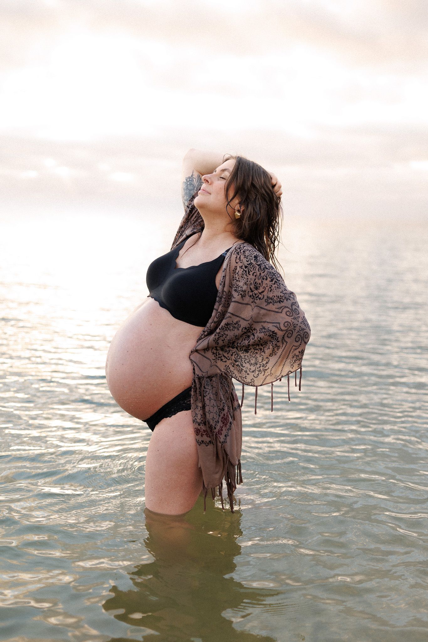 A pregnant woman wearing a shawl stands waist-deep in a body of water, enjoying the serene moment.