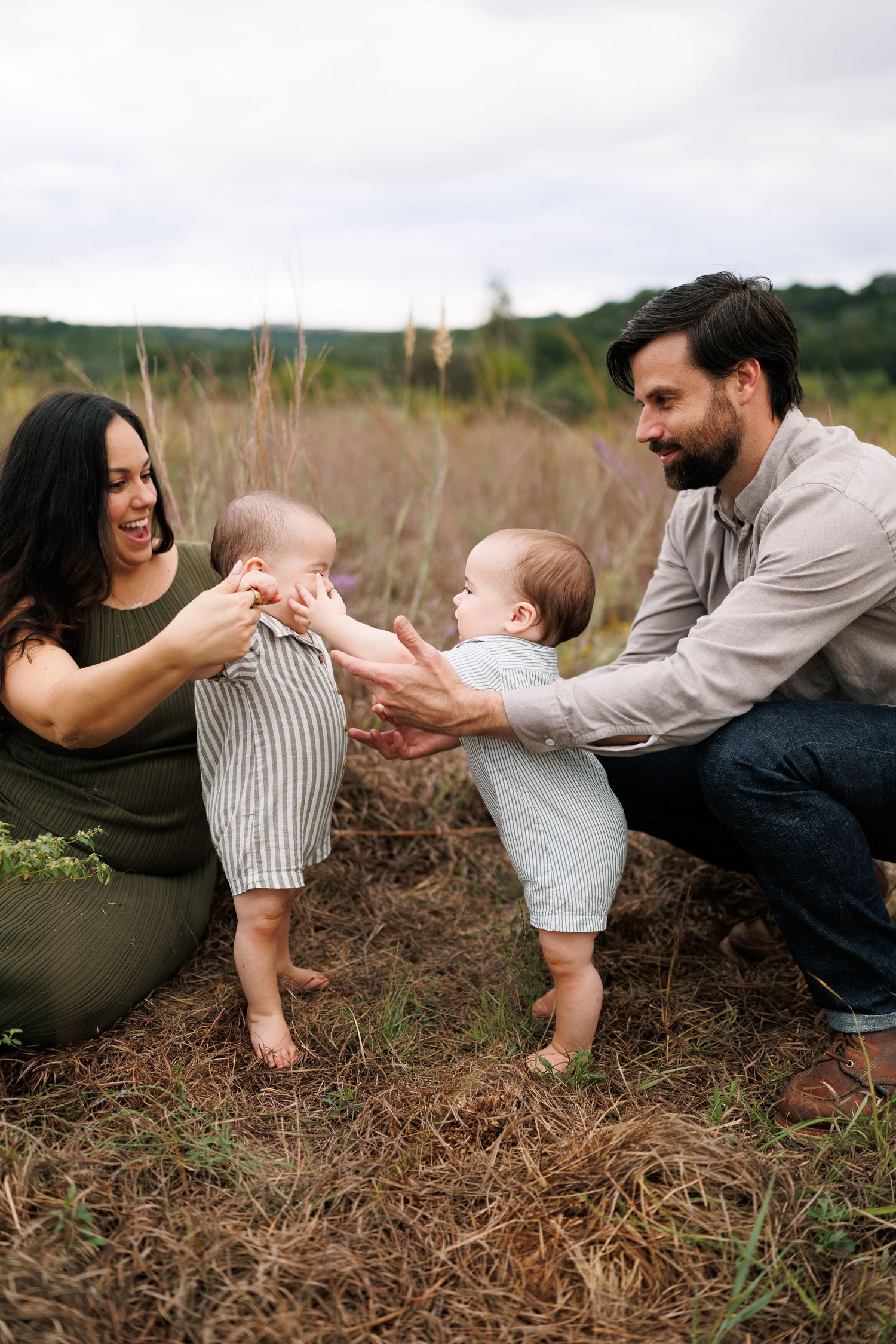 A happy family with two babies playing in a field, parents smiling as toddlers reach for each other outdoors.