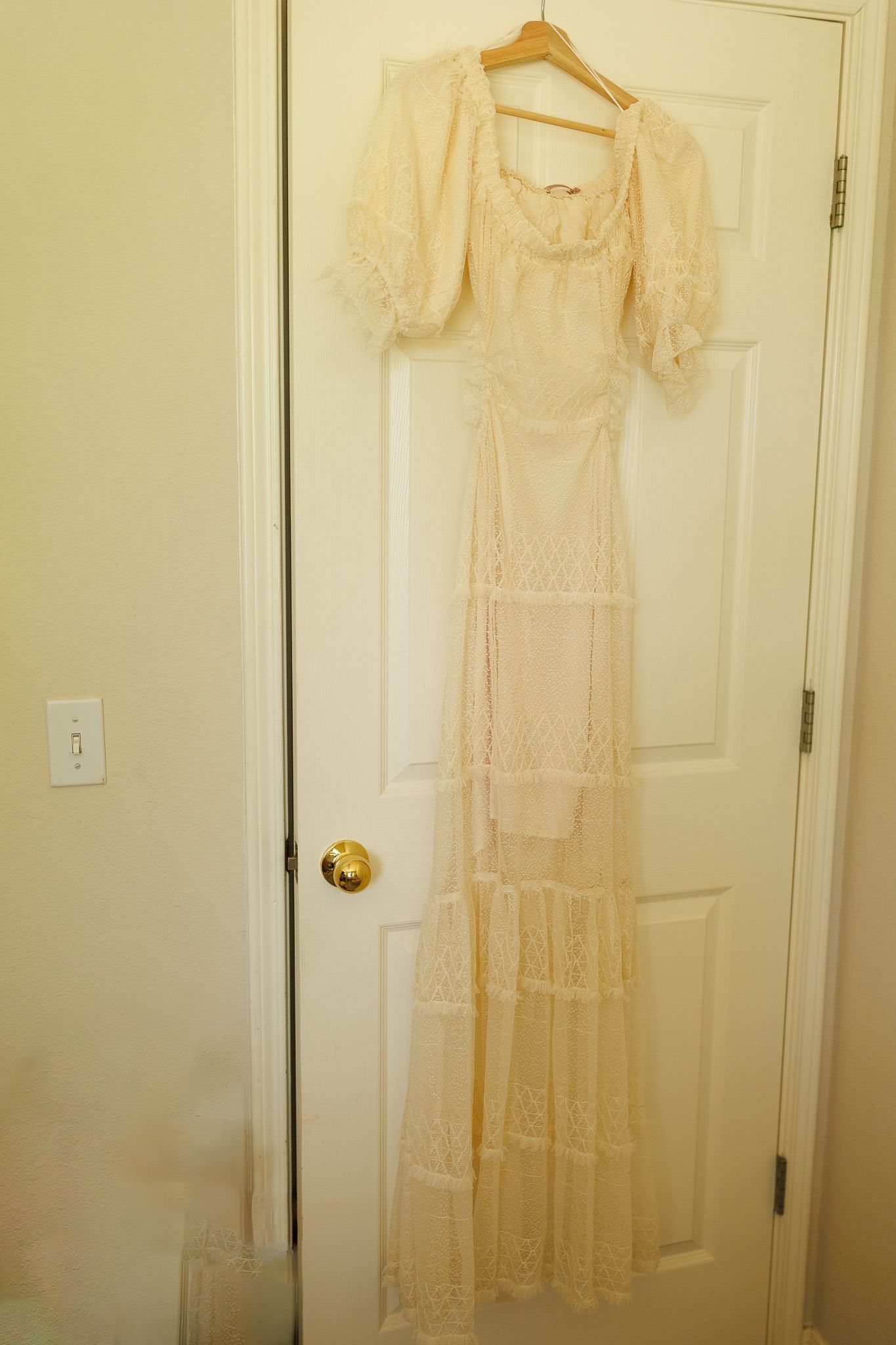 A vintage ivory lace dress hangs on a wooden hanger against a white door, displaying intricate floral embroidery patterns.