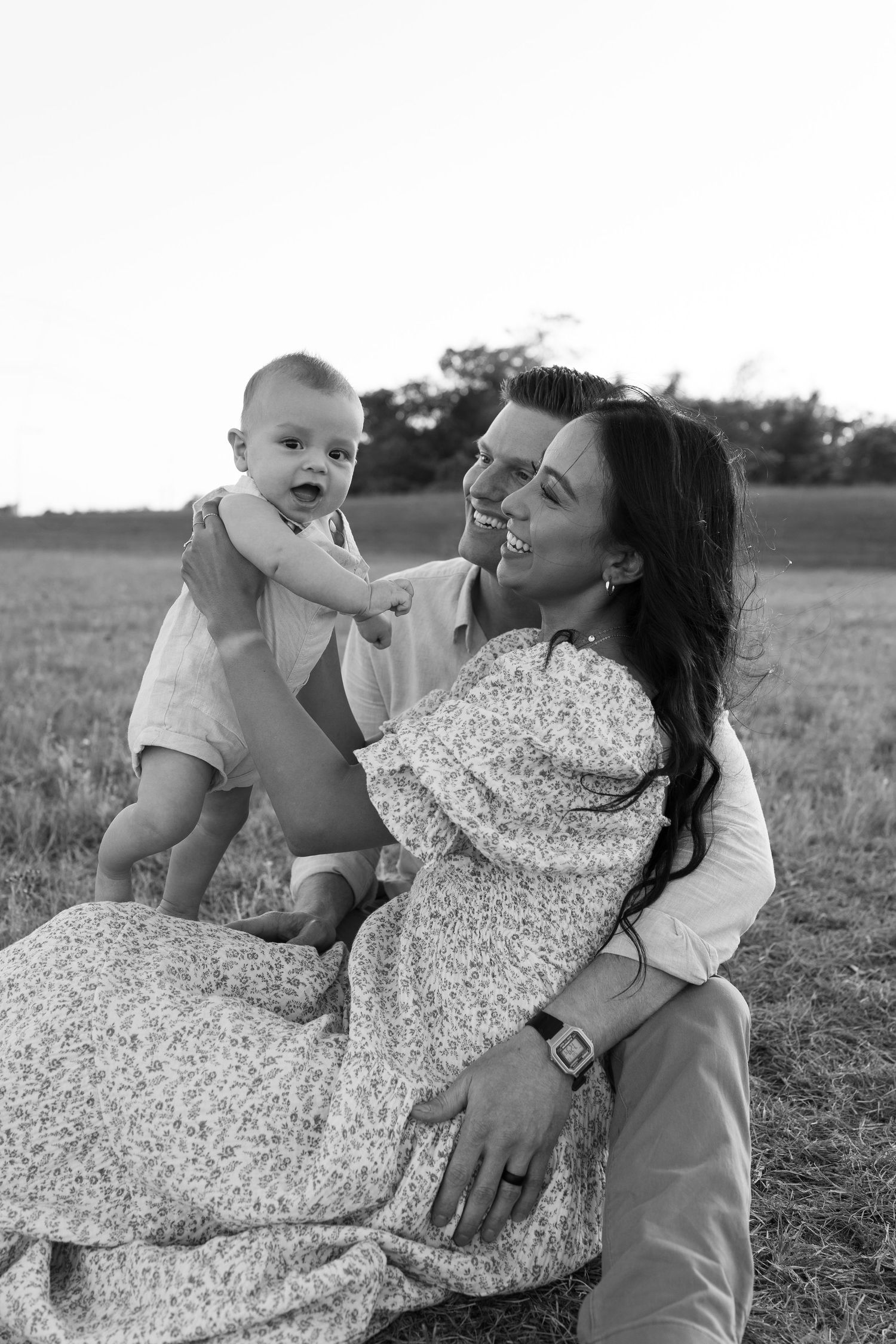 Black and white photo of a candid outdoor family moment with a baby and parents sitting together in a field.