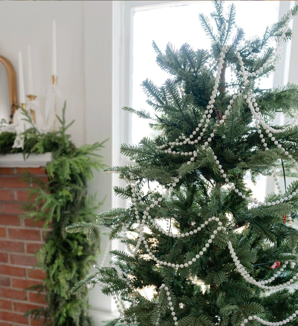 Christmas tree decorated with pearl garland strands near a brick fireplace mantel with greenery and candles.