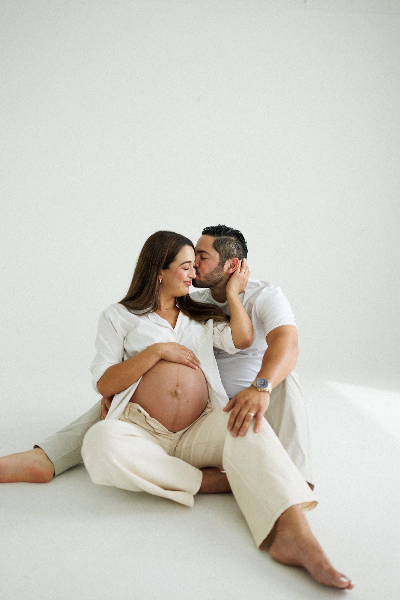 A pregnant woman and her partner sit on a white floor, smiling as he kisses her cheek while she cradles her baby bump.