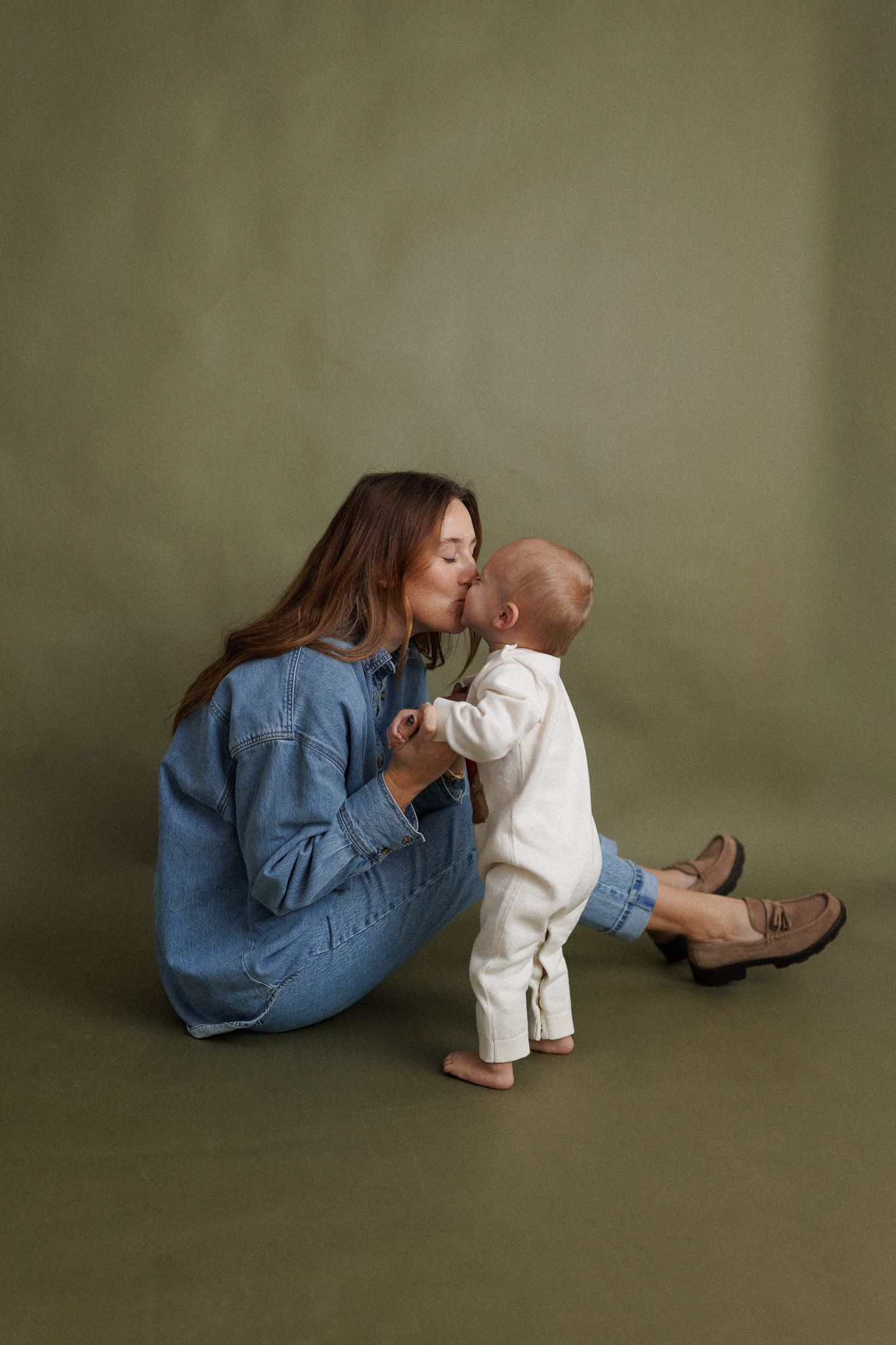 A tender moment between parent and baby in white outfit crawling on olive green backdrop in studio setting.